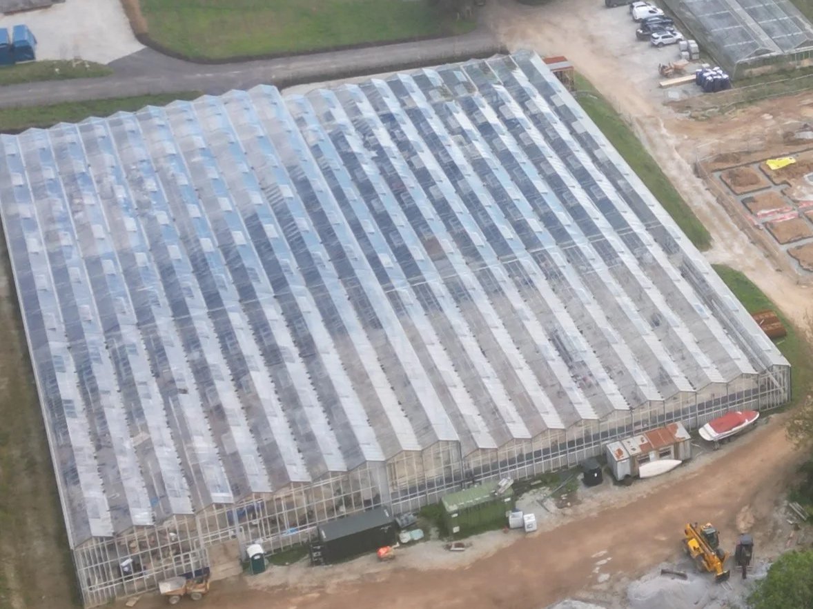 Aerial view of a large greenhouse with a glass roof, surrounded by green fields, dirt paths, and smaller buildings, with trees in the background.