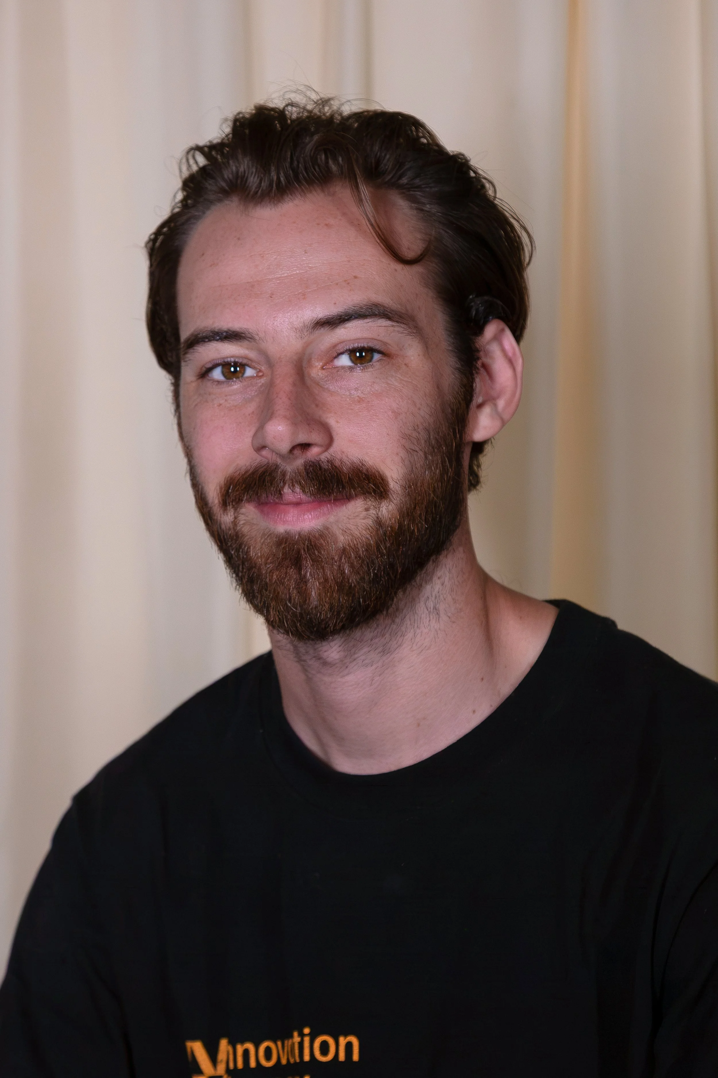 Close-up of a man with brown hair, brown eyes, a beard, and a mustache, wearing a black shirt with orange text on it, with a beige curtain in the background.