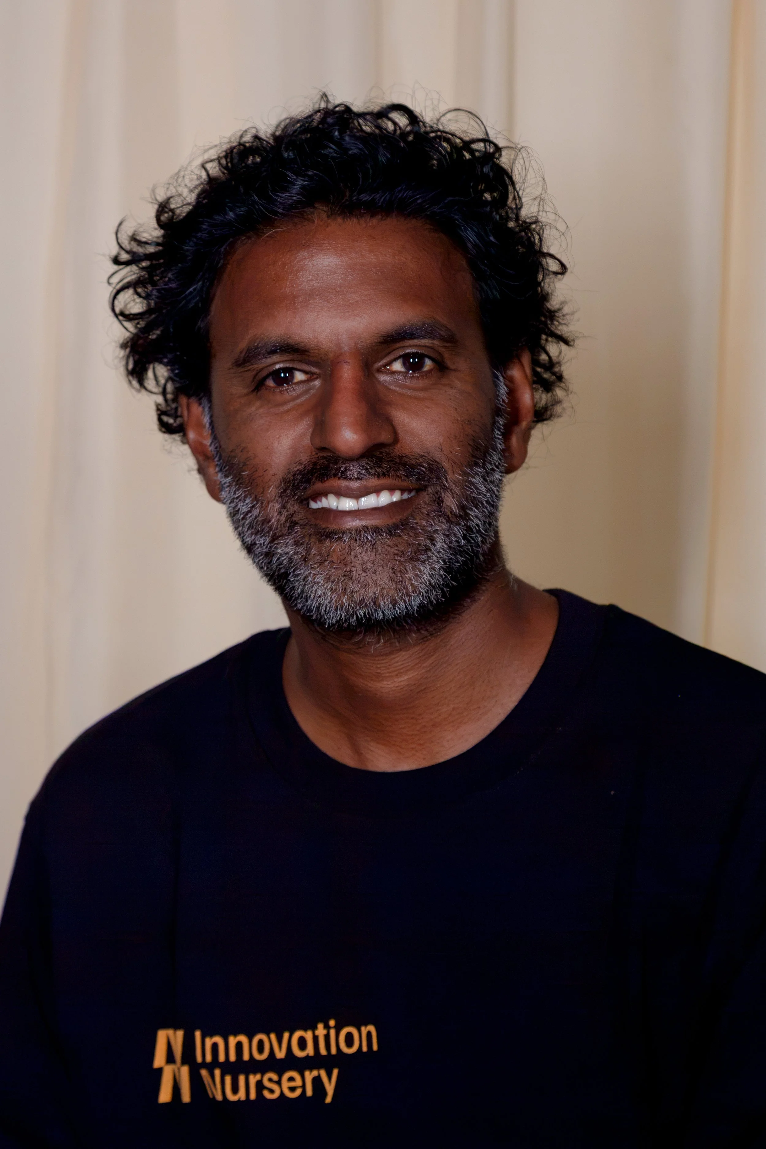 A man with dark curly hair and a beard, smiling, wearing a black shirt with orange text that reads 'Innovation Nursery', standing in front of a light-colored wall.
