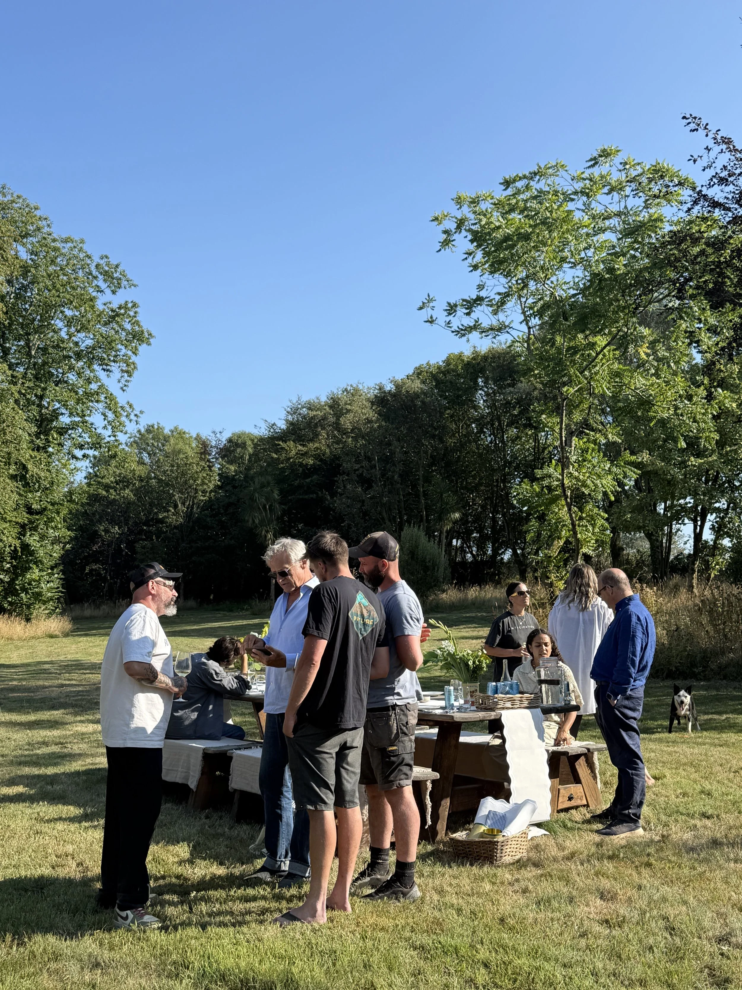 People gathered outdoors on our site, around a table with food and drinks, with trees and a clear blue sky in the background.