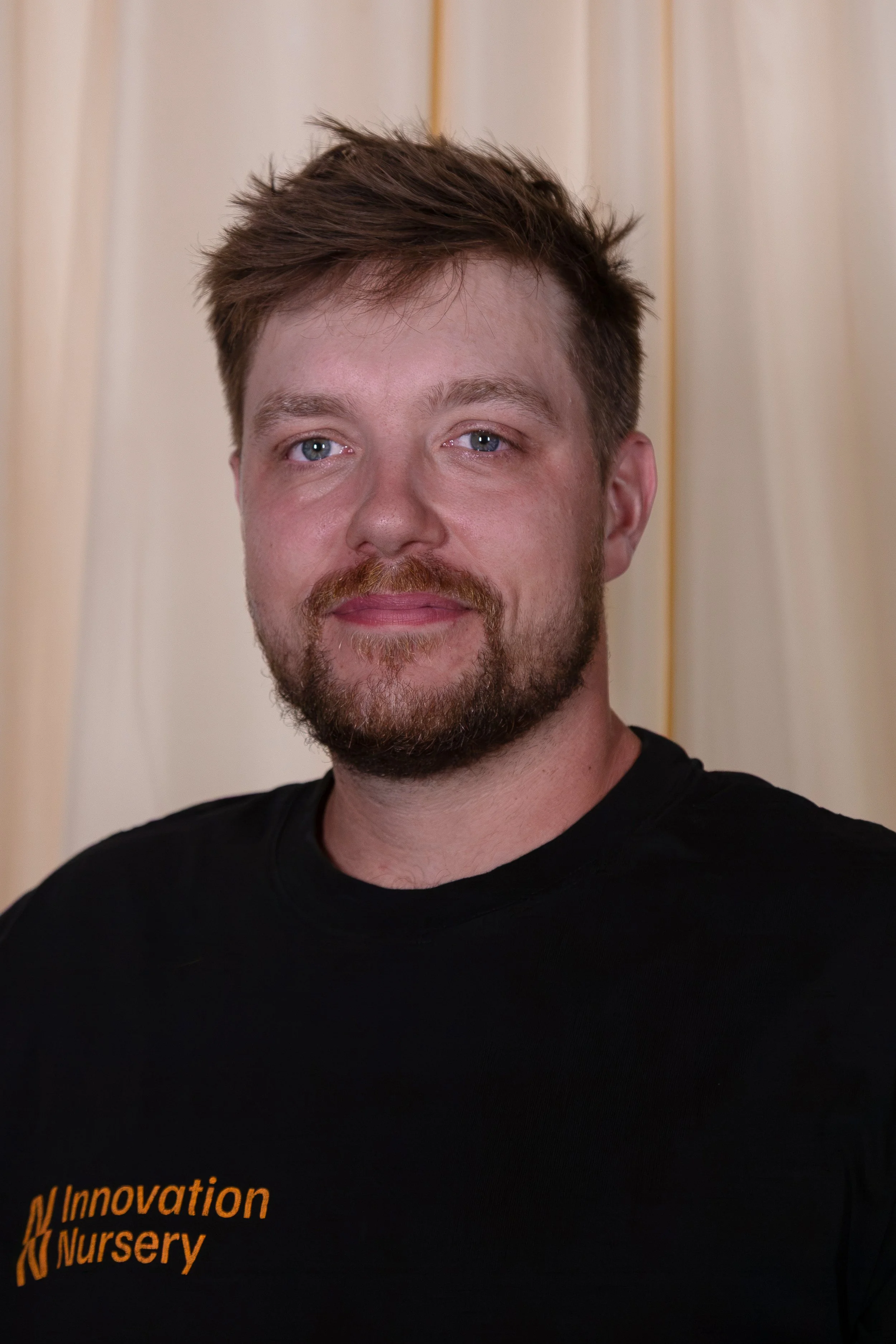 Close-up of a man with light skin, brown hair, and a beard, wearing a black T-shirt with orange text that says 'Innovation Nursery,' standing against a beige curtain background.