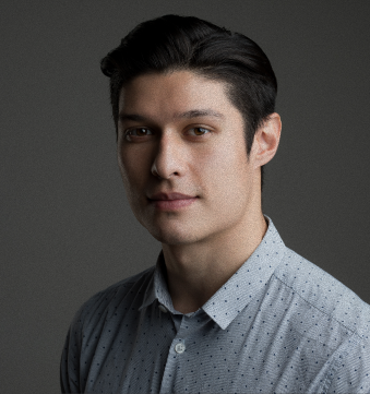 A young man with dark hair and fair skin wearing a light blue, button-up shirt with small dark patterns, against a dark gray background.