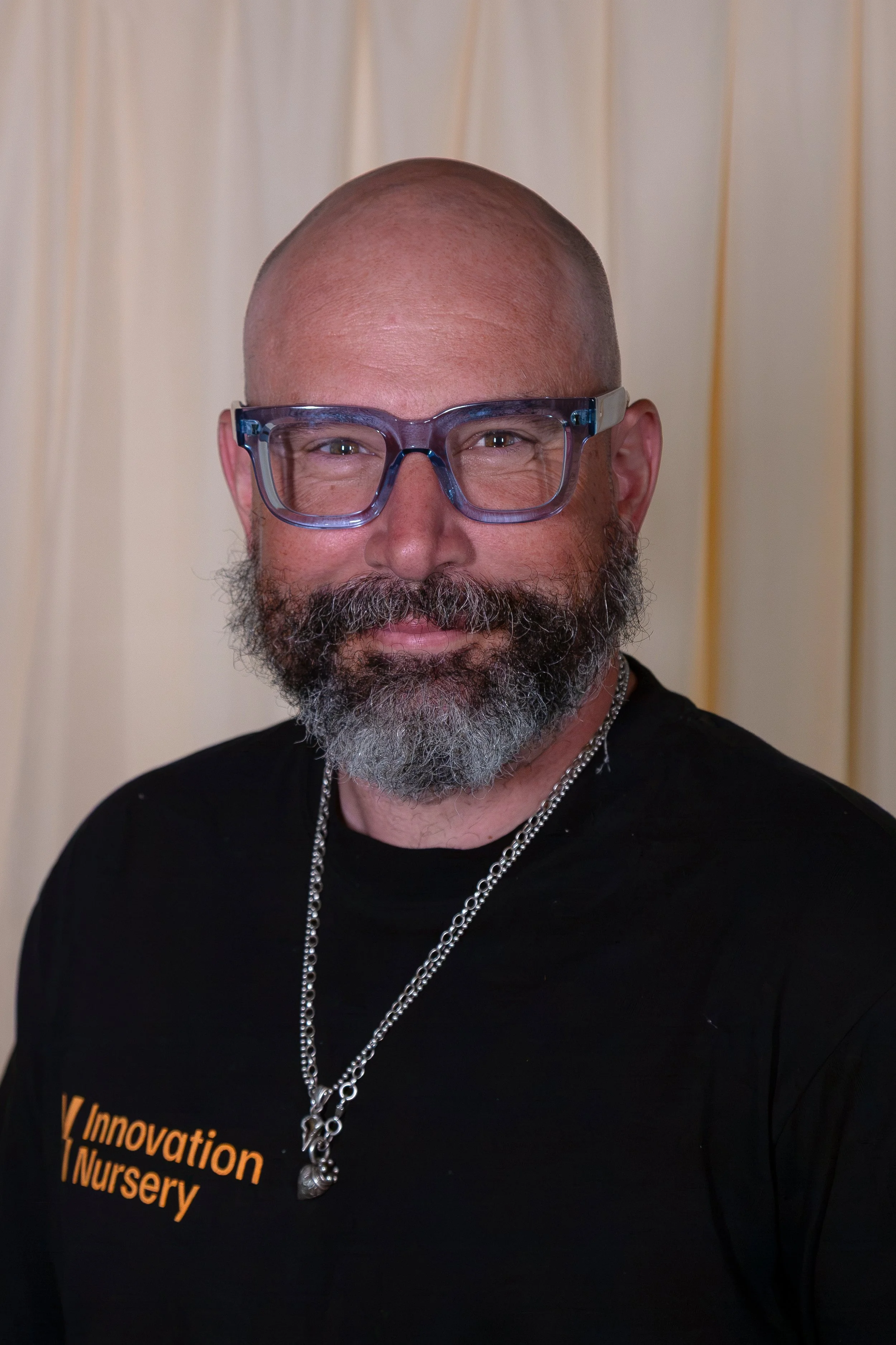 A bald man with glasses and a beard smiling, wearing a black shirt with orange text that says 'Innovation Nursery' and two silver necklaces, standing in front of a beige background with vertical lines.