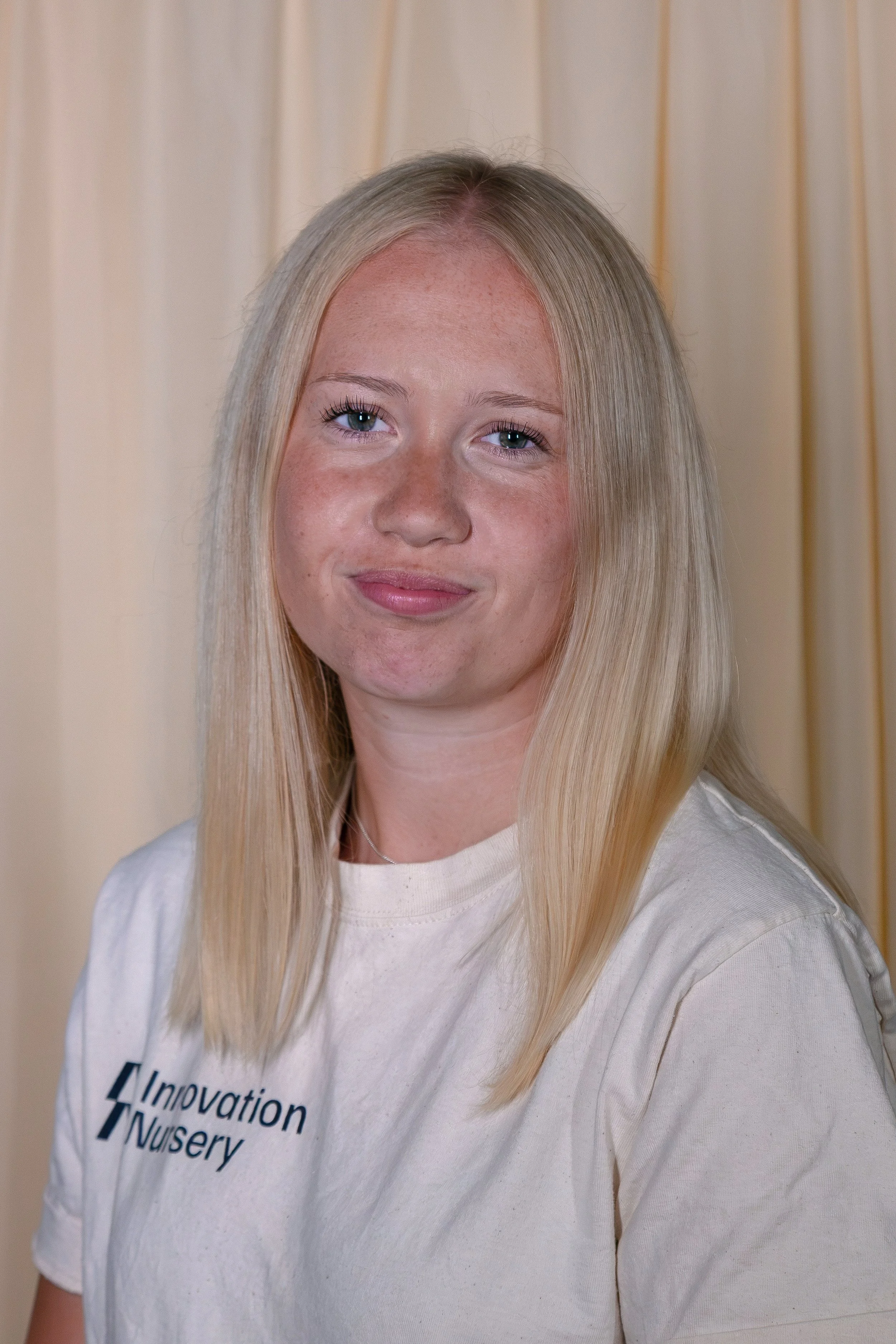 A young woman with long blonde hair and blue eyes, wearing a white T-shirt with text on it, standing against a beige curtain background.