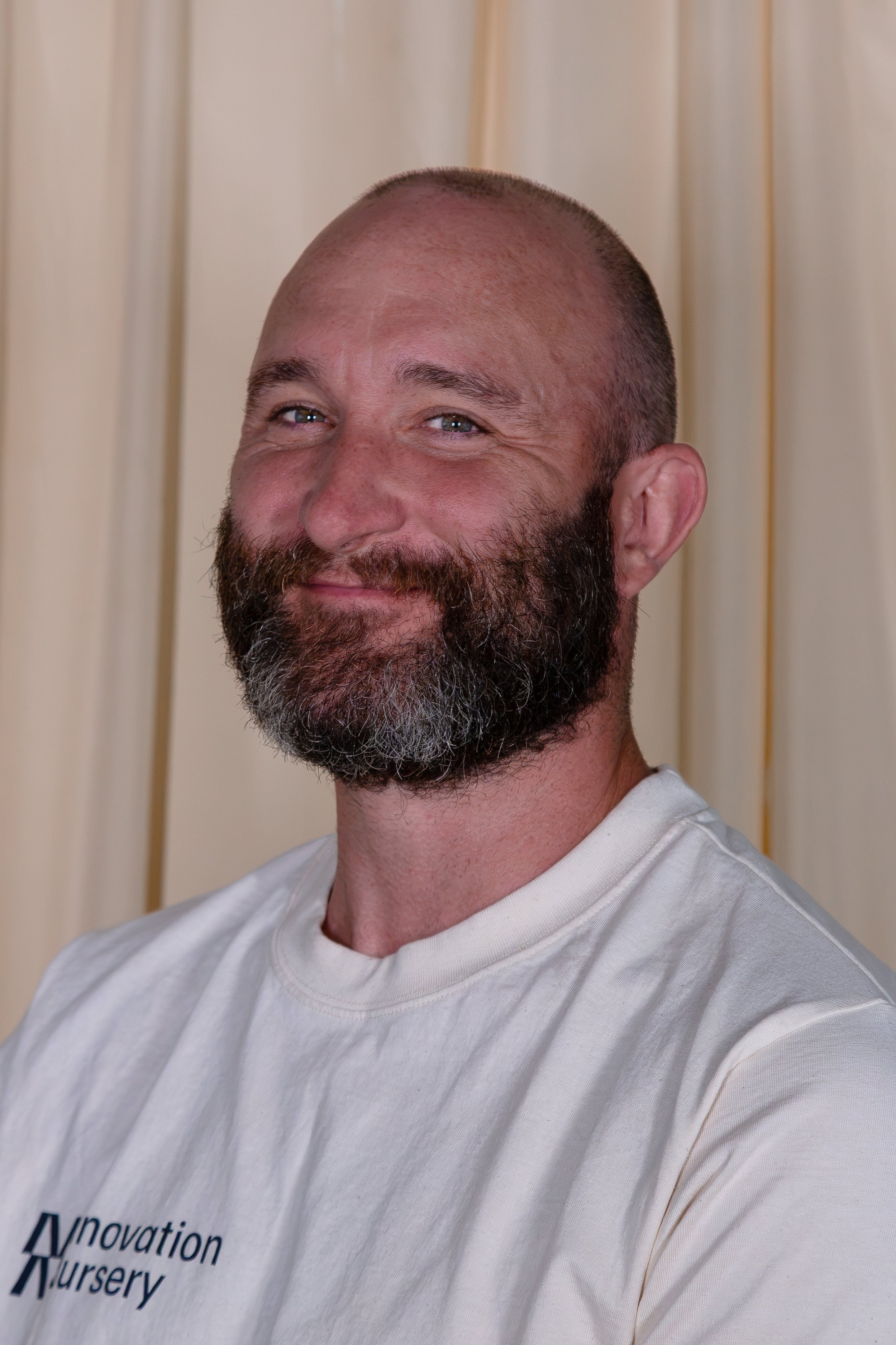 Close-up of a smiling man with a beard and shaved head wearing a white shirt with 'Innovation Nursery' logo, standing in front of beige curtains.