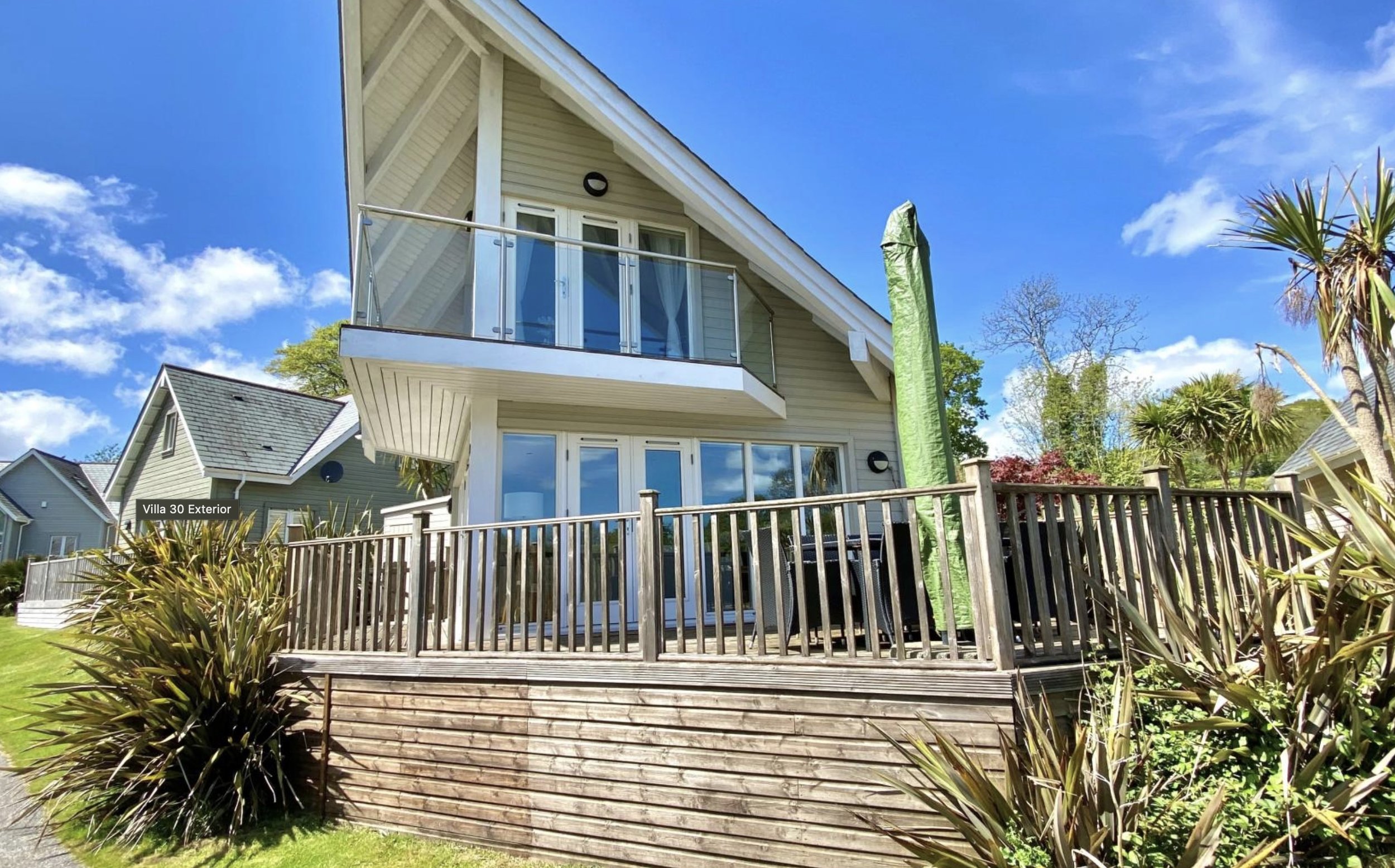 Exterior view of a modern two-story house with a wooden deck, sliding glass doors, and a glass balcony railing, surrounded by greenery and palm trees under a partly cloudy blue sky.