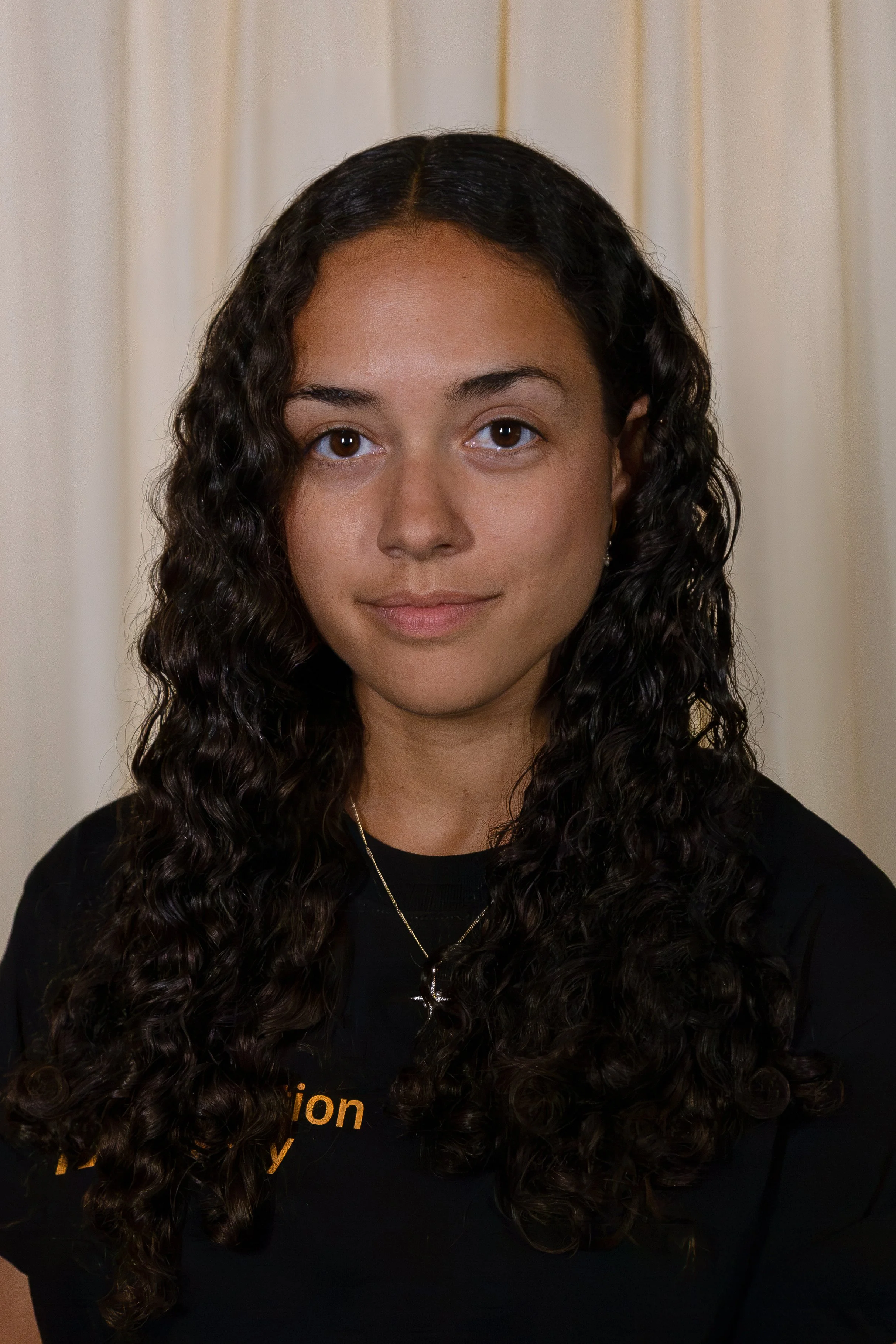 A woman with long, curly dark hair, wearing a black top and a gold necklace, standing in front of a light-colored curtain.