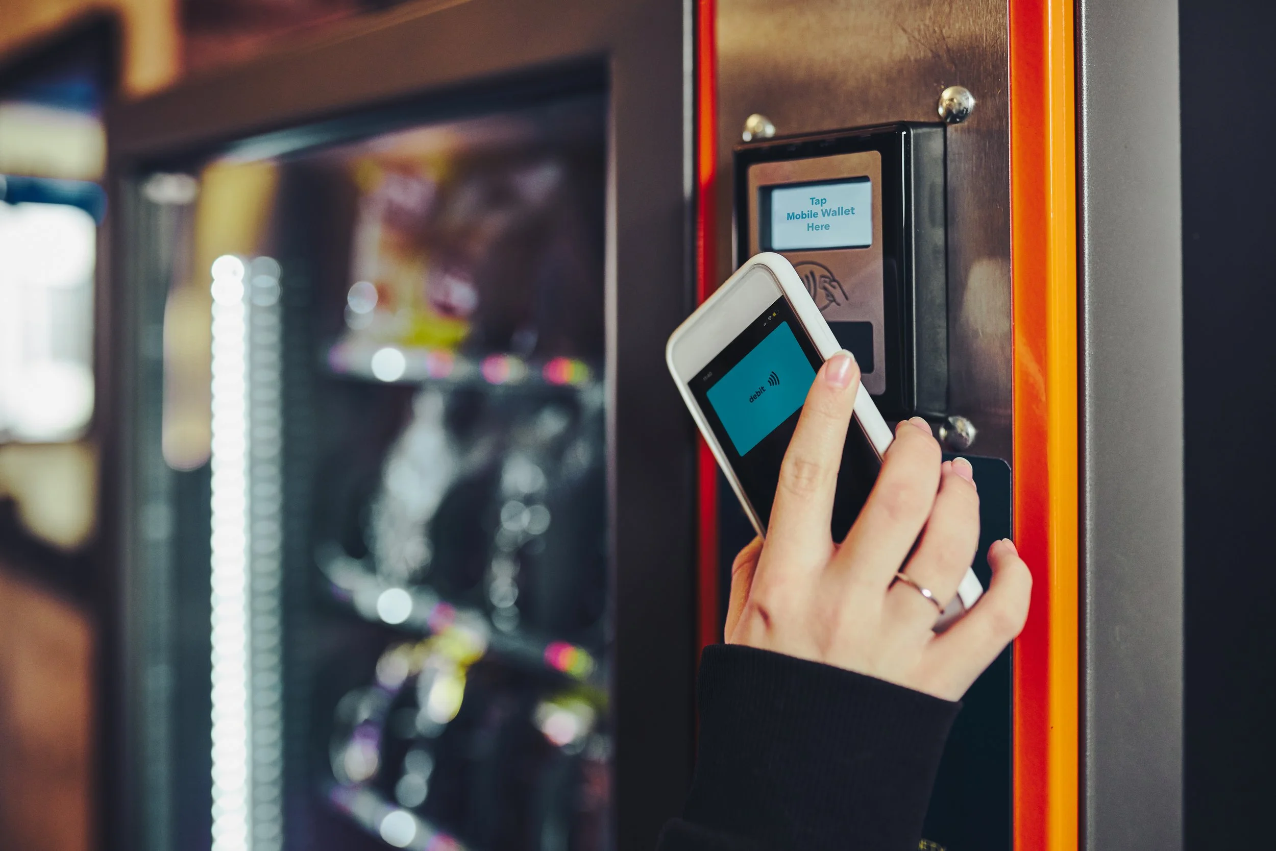 Person holding a smartphone near a vending machine with a screen displaying 'Tap Mobile Wallet Here'.