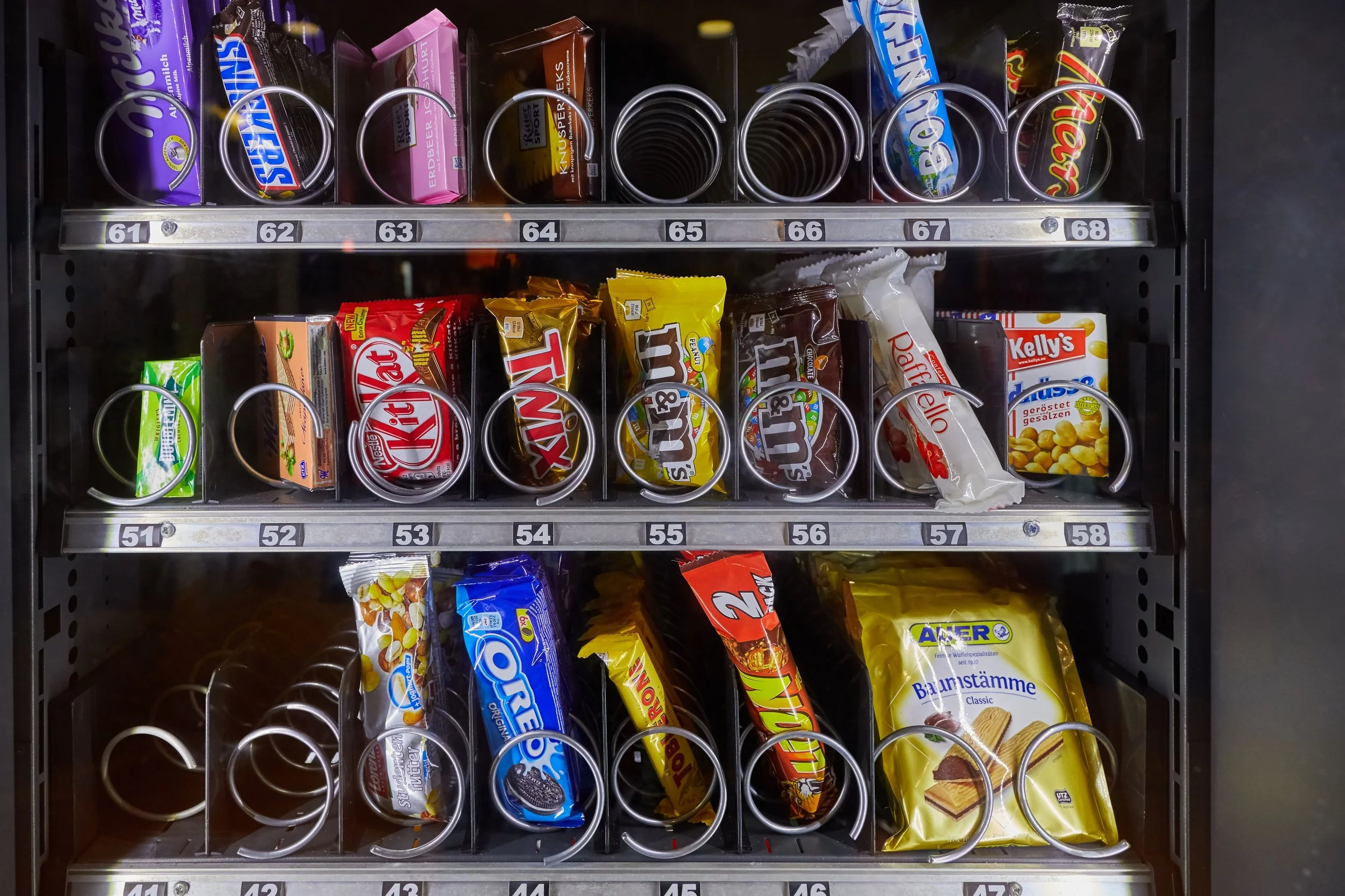Vending machine containing various snack bars, chocolates, and cookies arranged in rows and slots, including brands like Snickers, KitKat, Bounty, and Kelly's.