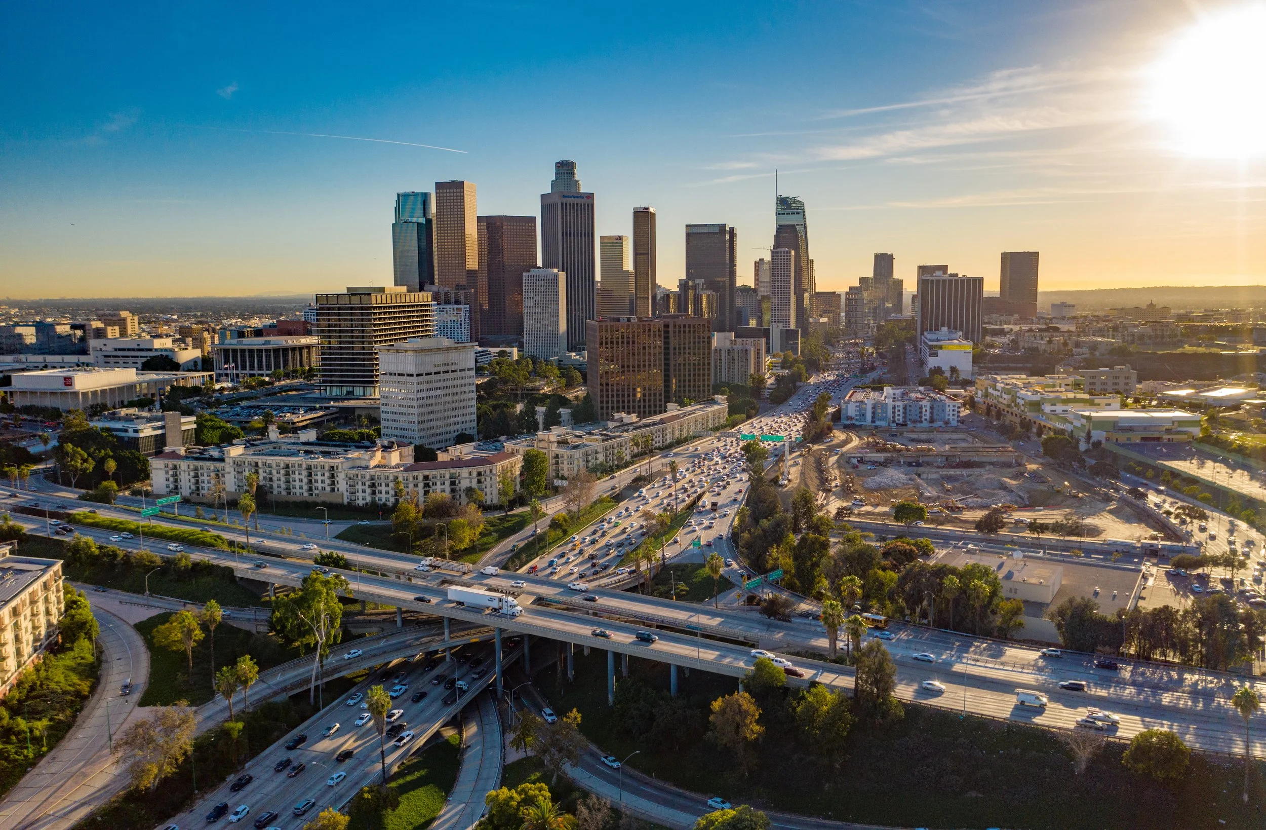 Los Angeles Orange County Inland Empire skyline with tall buildings and skyscrapers at sunset, with highways and traffic in the foreground and a clear sky.