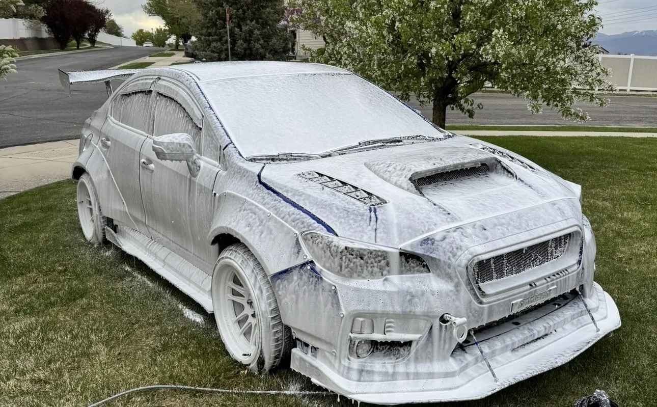 A silver car covered in soap suds and foam during a car wash in a suburban neighborhood, with trees, houses, and a mountain range in the background.