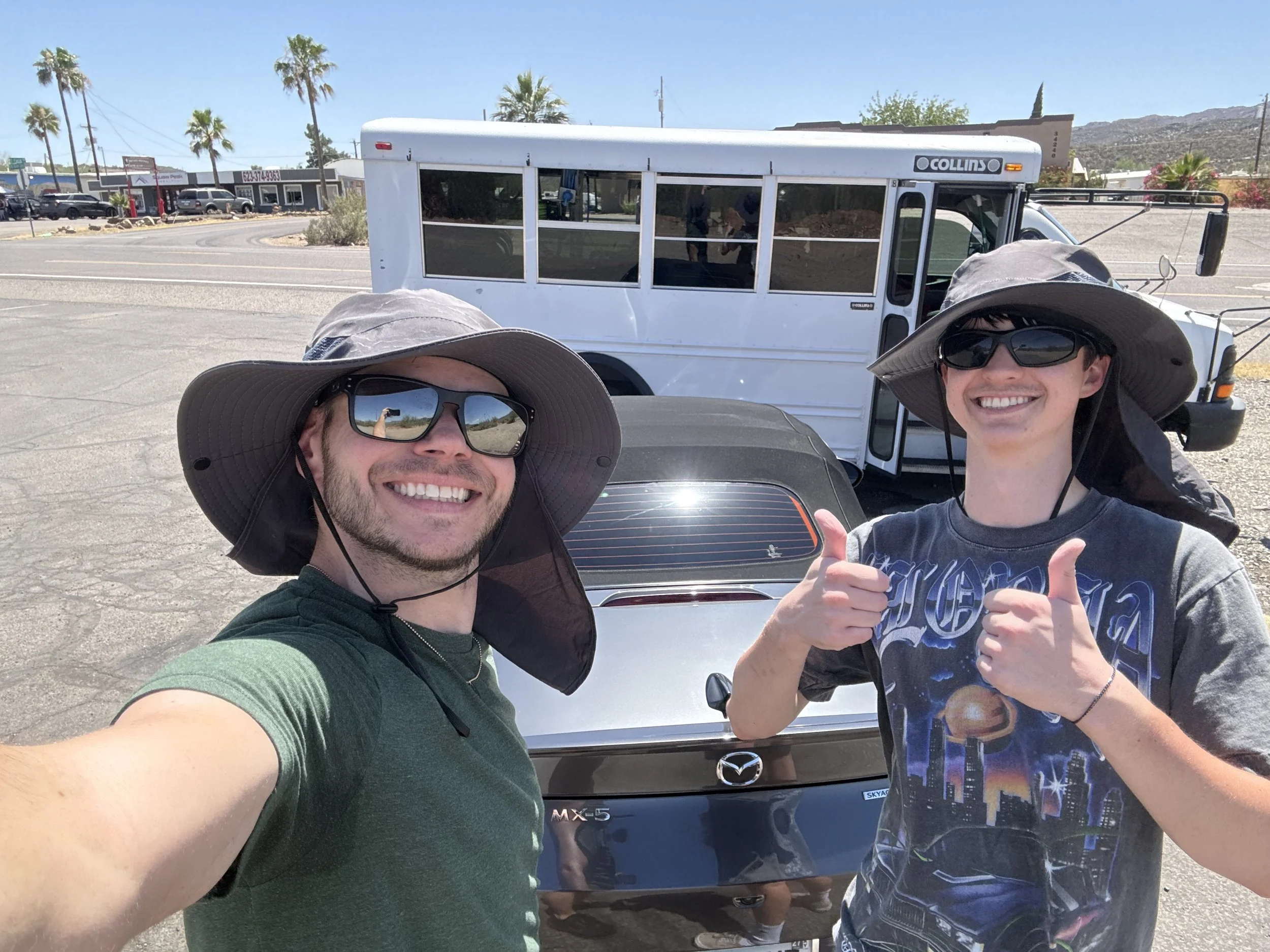 Two smiling young men wearing wide-brimmed hats and sunglasses taking a selfie in front of a white shuttle bus on a sunny day. One man is giving a thumbs-up gesture.
