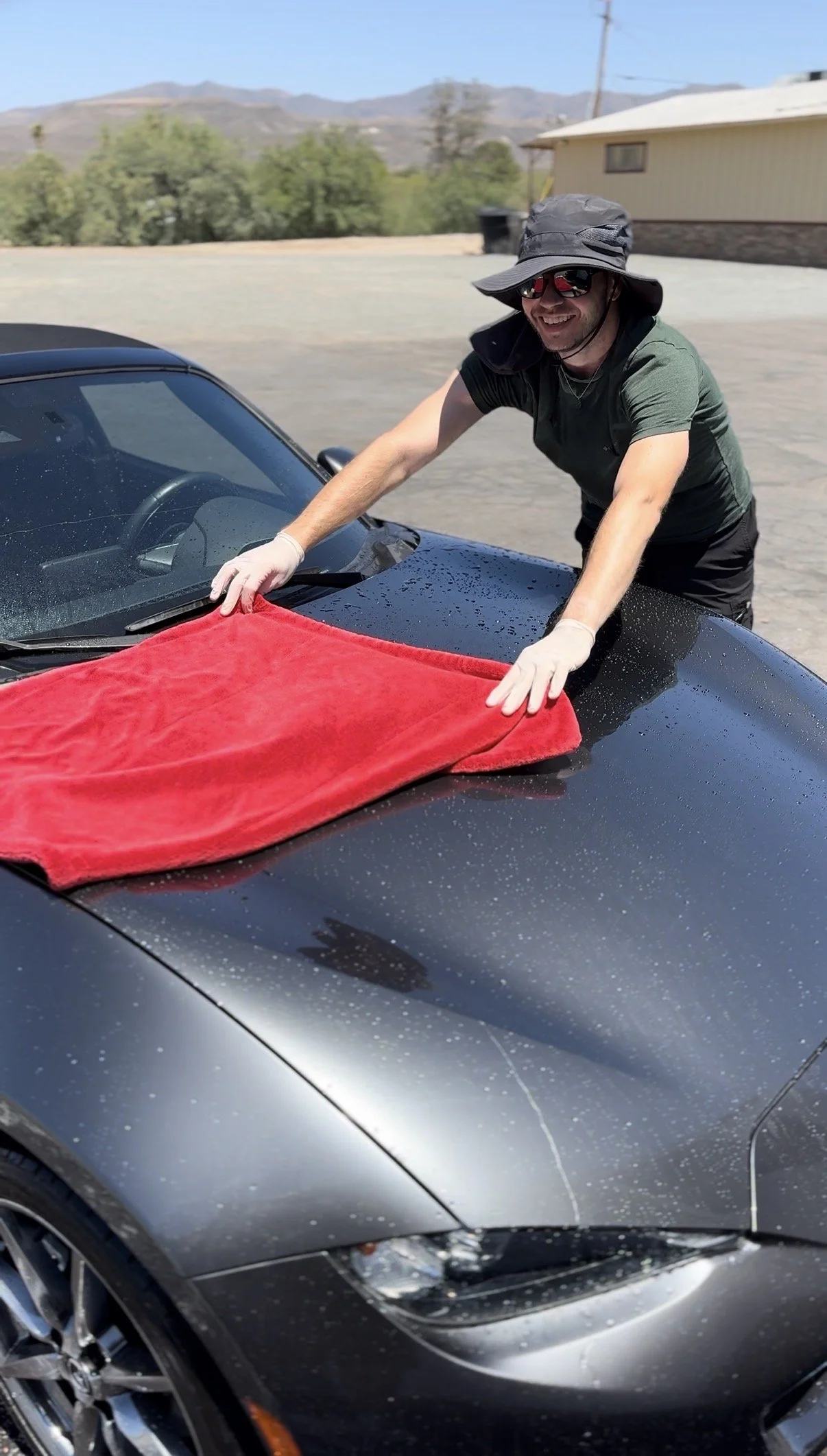 A man wearing a hat, sunglasses, and gloves is cleaning the hood of a black sports car with a red towel. The car is wet, and the background shows a house, trees, and mountains.