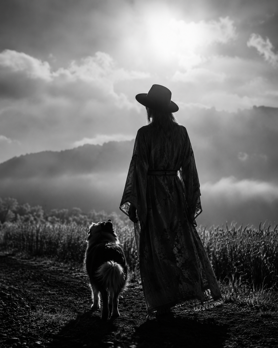 A black-and-white photo of a woman wearing a wide-brimmed hat and a long dress, walking with a dog along a trail in a scenic outdoor landscape with mountains and clouds in the background.
