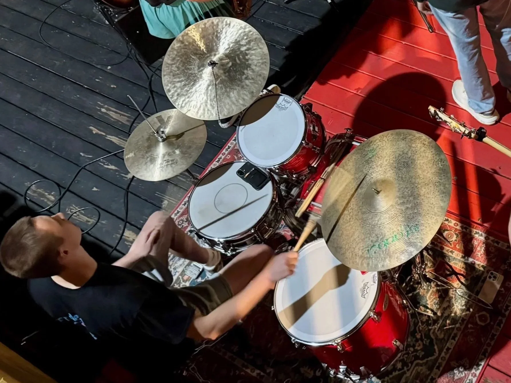 Isaiah Vinay playing drums from a top-down angle during a live music performance, with cymbals, a red drum kit, and stage gear around him.