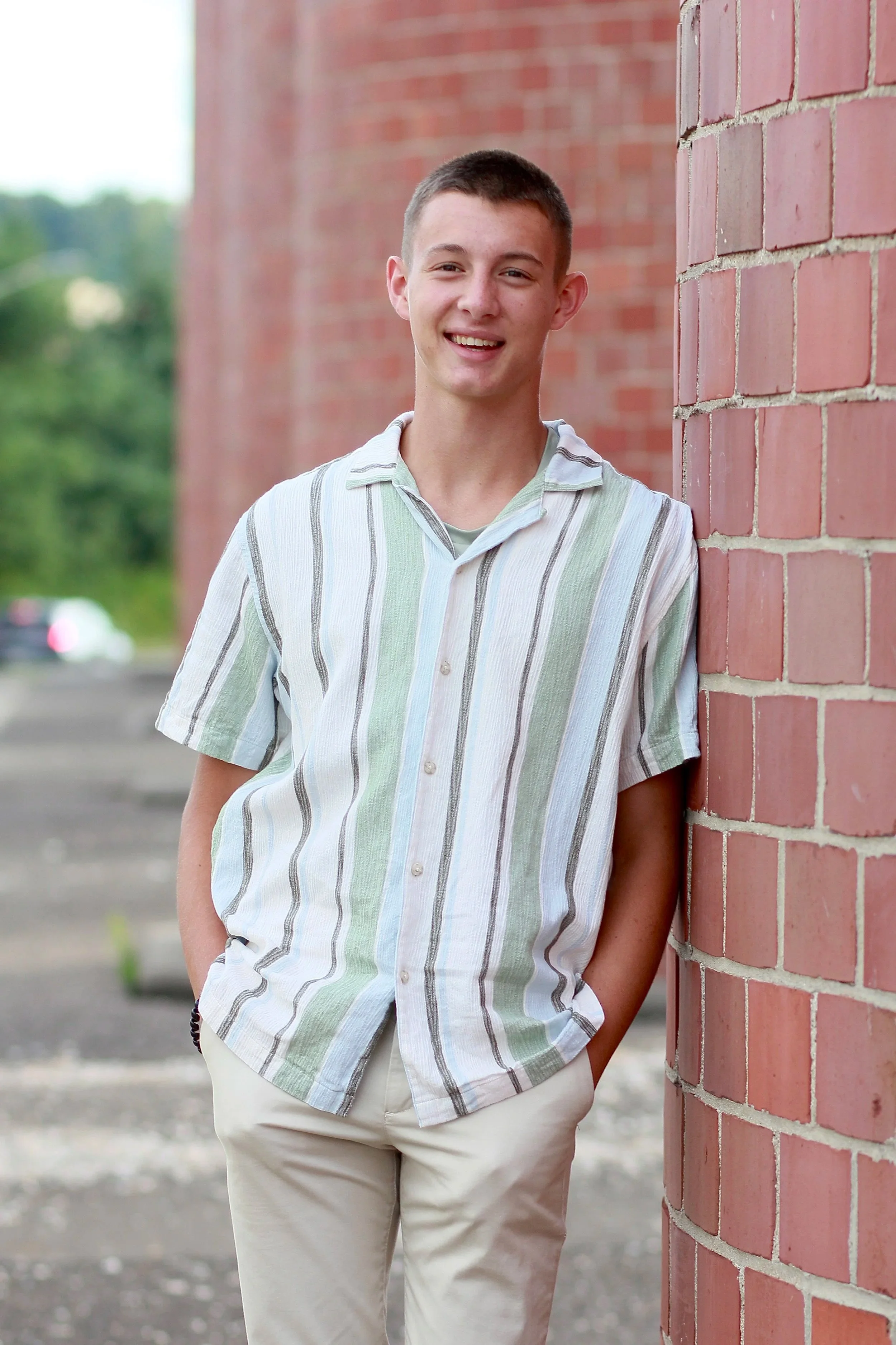 Isaiah Vinay standing outdoors beside a red brick wall, wearing a striped button-up shirt and light-colored pants.