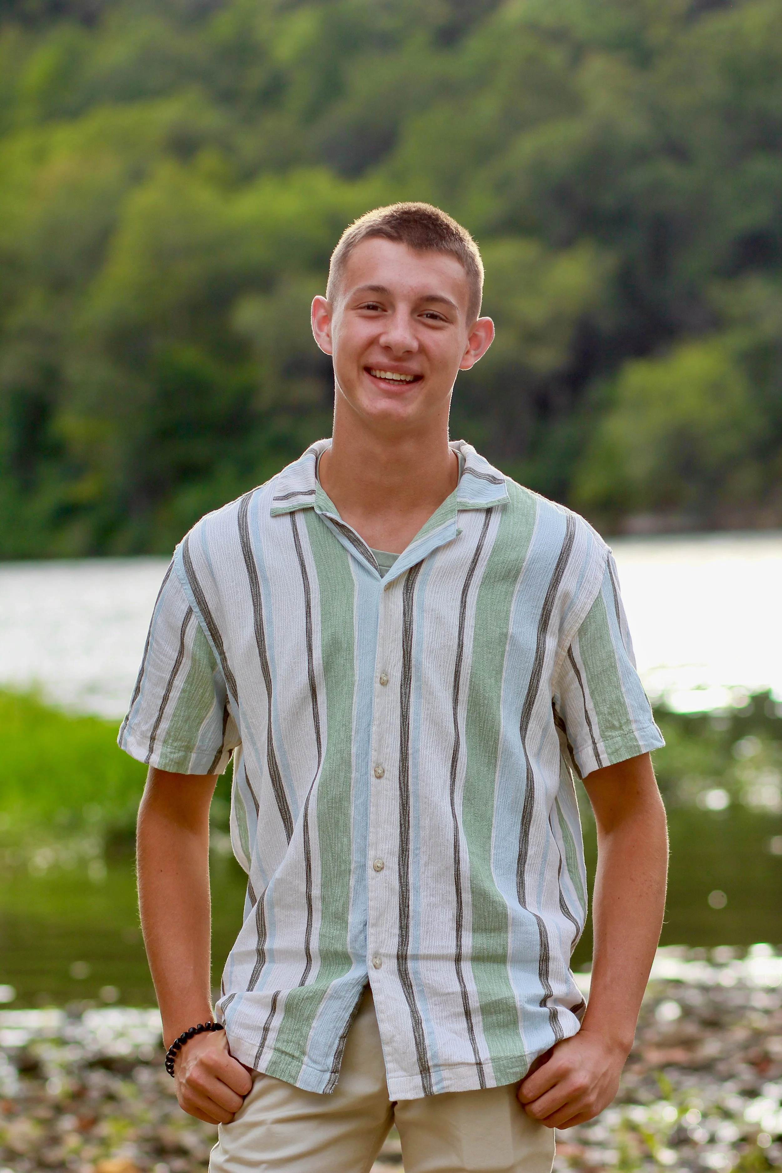Isaiah Vinay standing outdoors near water and trees, wearing a striped shirt and smiling for an About page portrait.