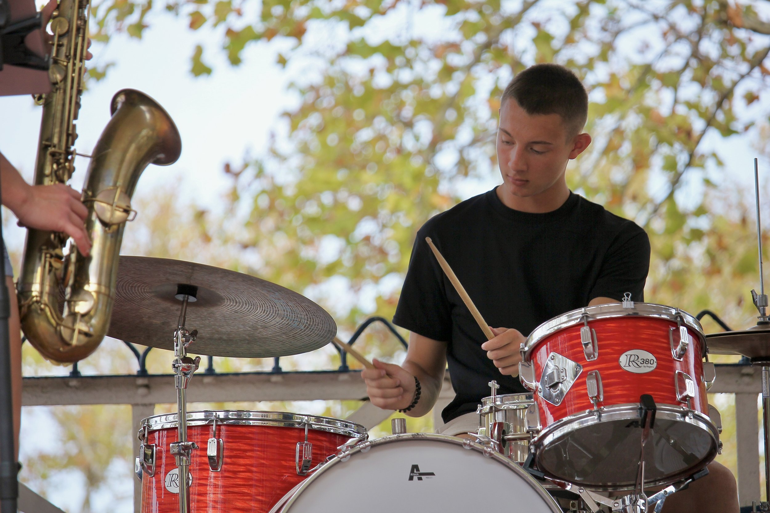 Isaiah Vinay playing drums during an outdoor live music performance with saxophone and band members nearby.