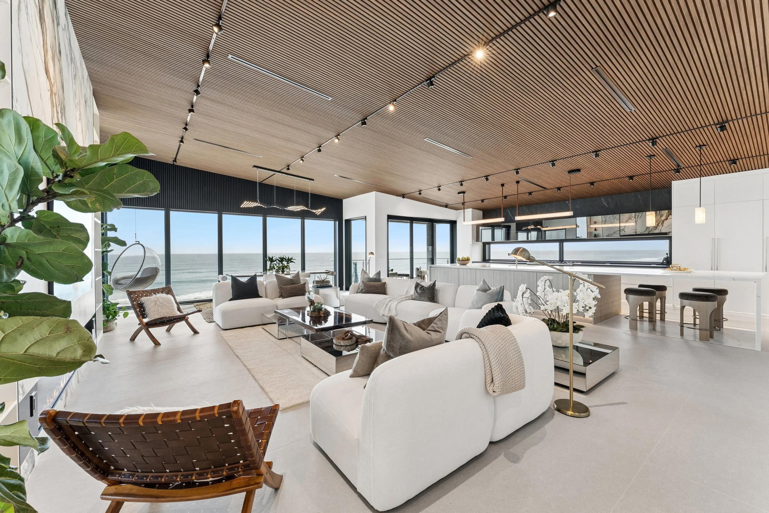 Modern living room with large windows overlooking the ocean, white sofas, a wooden chair, green plants, and stylish lighting on a wooden ceiling.