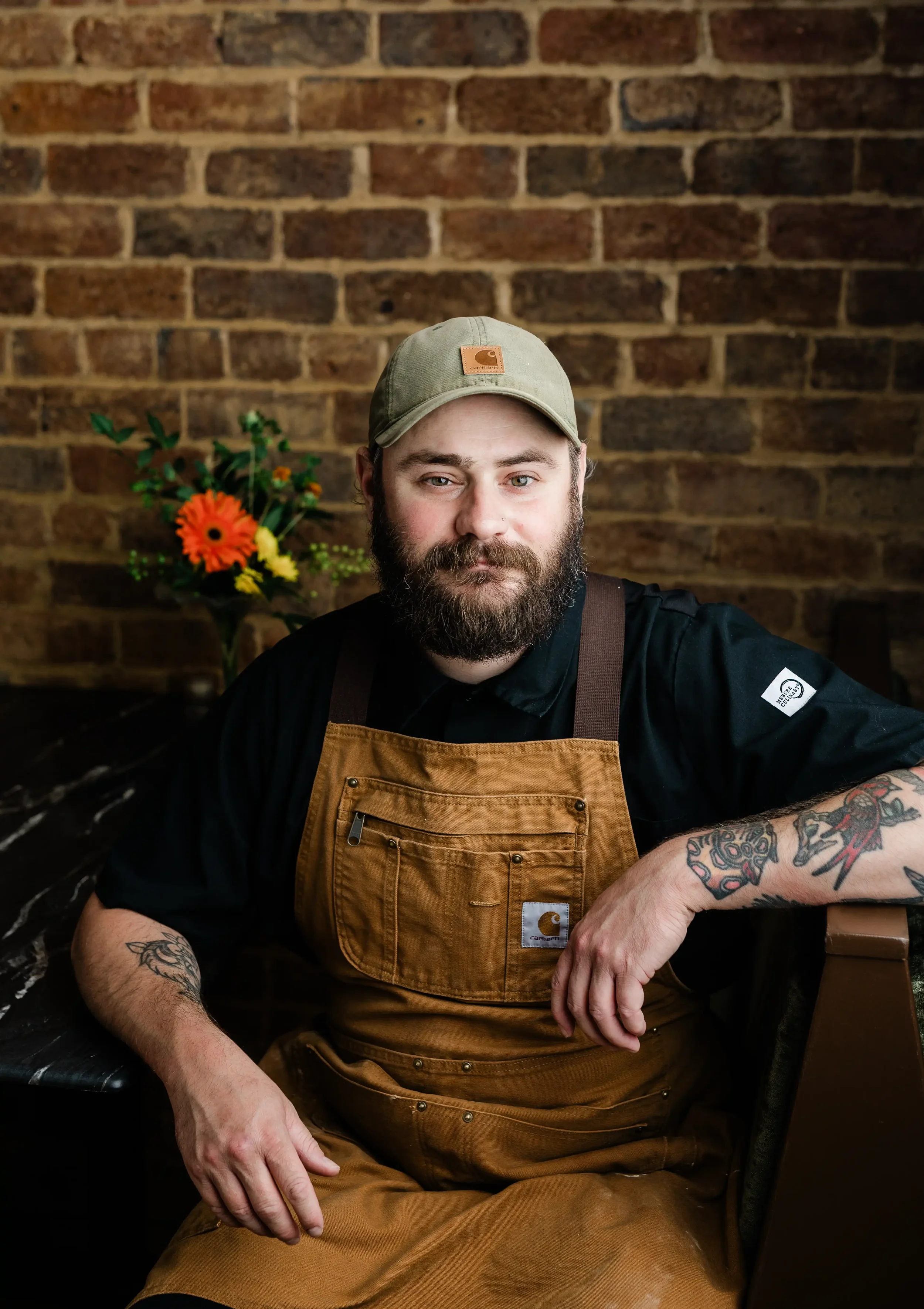 A bearded man wearing a tan Carhartt cap and a brown work apron, sitting in front of a brick wall with a bouquet of colorful flowers in the background.