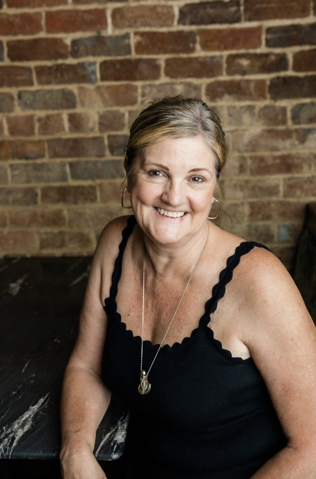 A smiling woman with blonde wavy hair, wearing a beige top, a floral kimono, and a dragon pendant necklace, sitting in front of a brick wall with a plant to her left.