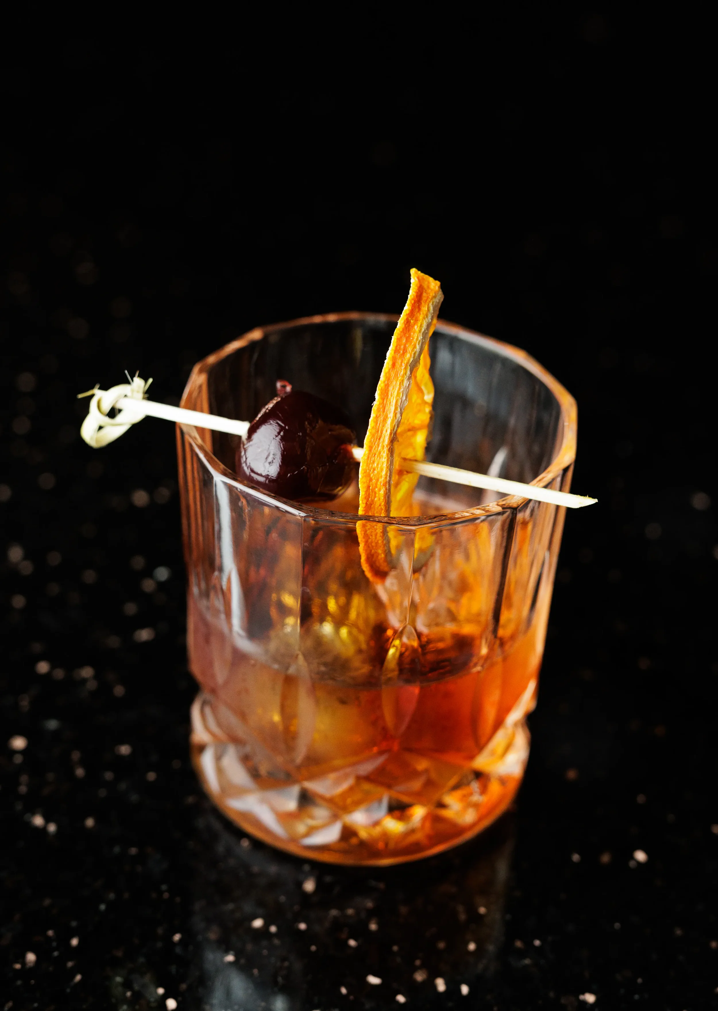 A close-up of a cocktail glass with amber-colored drink, garnished with a cherry on a skewer and a twist of orange peel, set against a dark, speckled background.