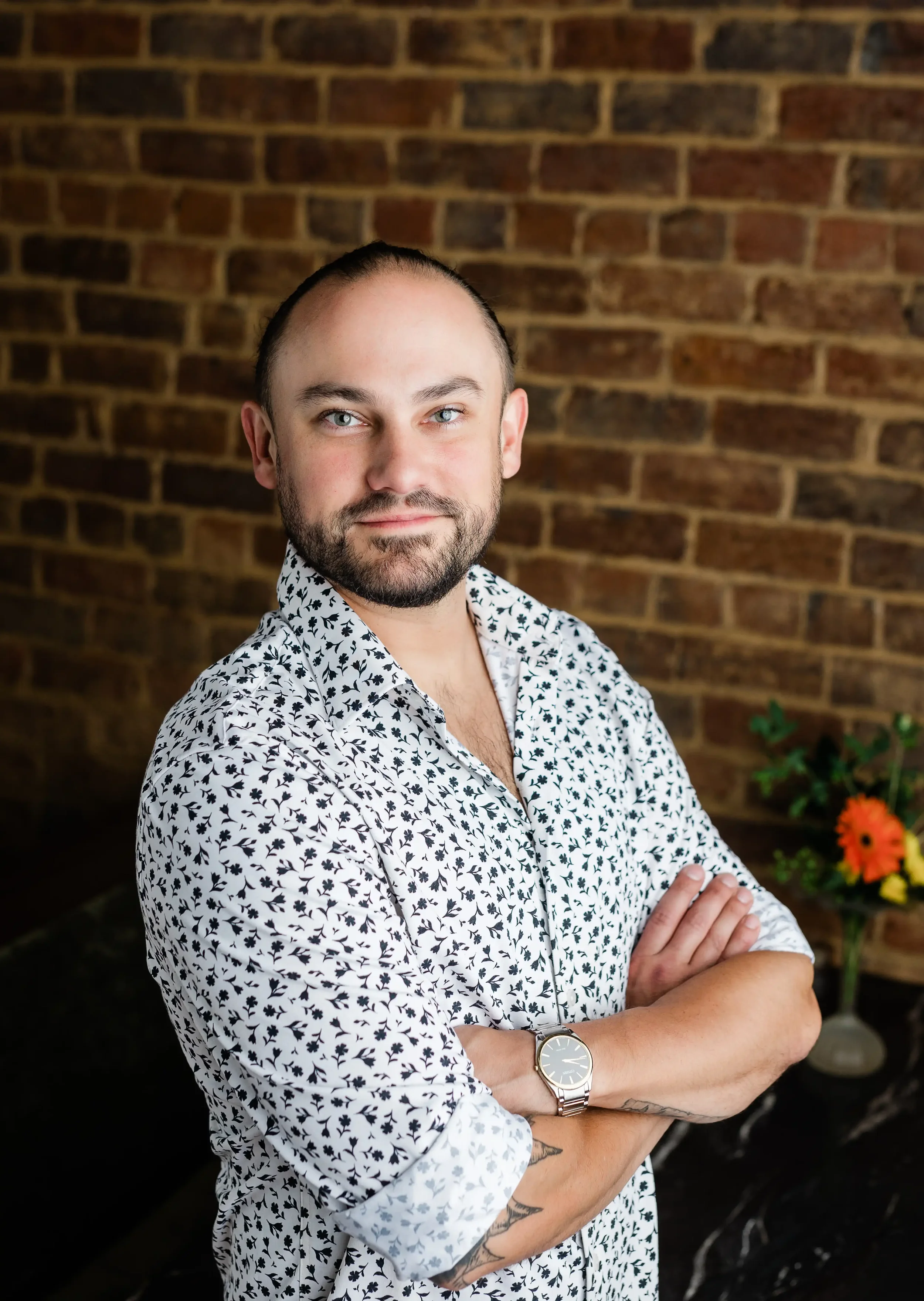 A man with light skin, blue eyes, and a beard, standing with arms crossed in front of a brick wall, wearing a white shirt with black leaf patterns and a watch on his left wrist.