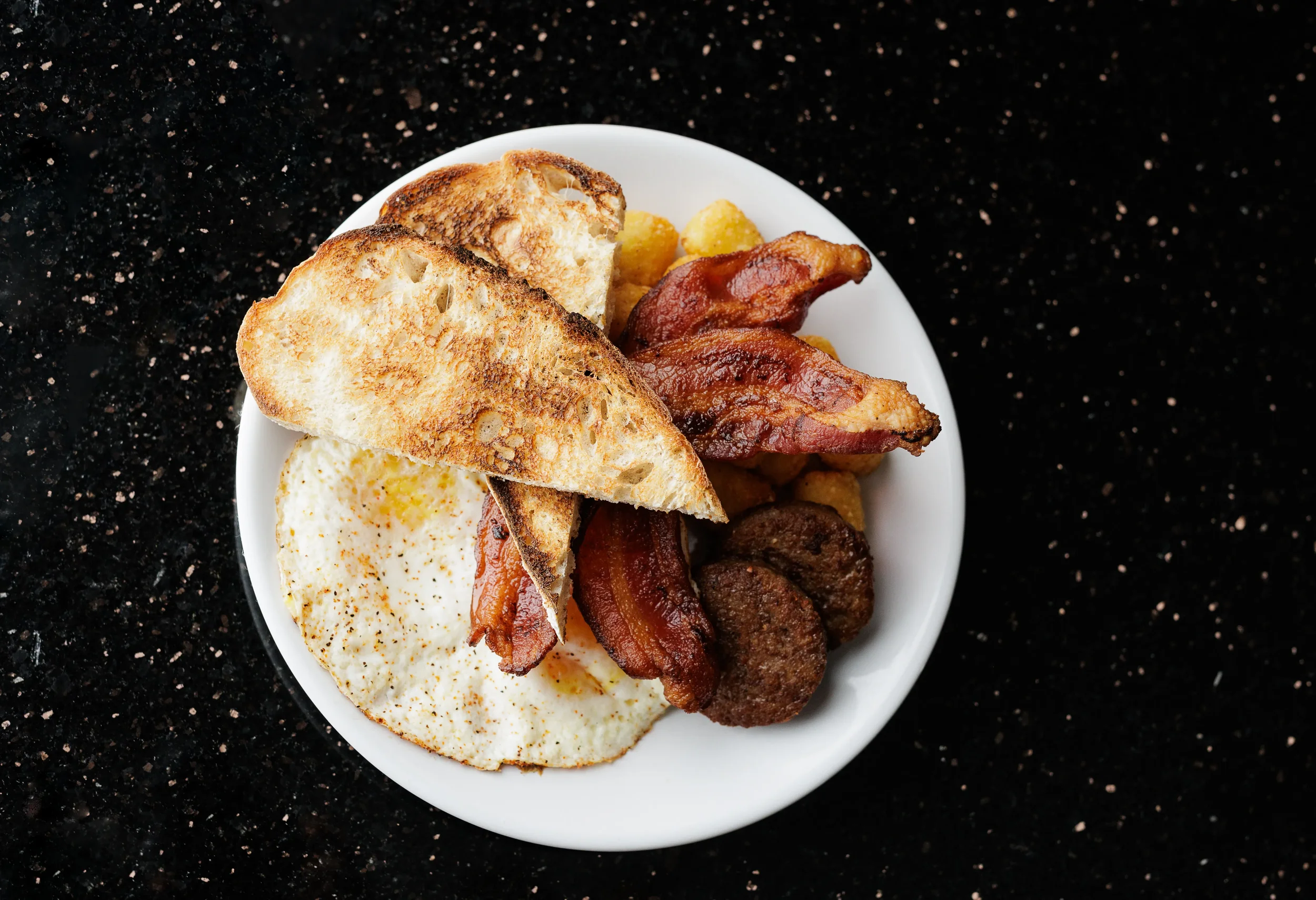 A white plate containing toast, fried eggs, bacon, sausage patties, and breakfast potatoes on a black speckled countertop.