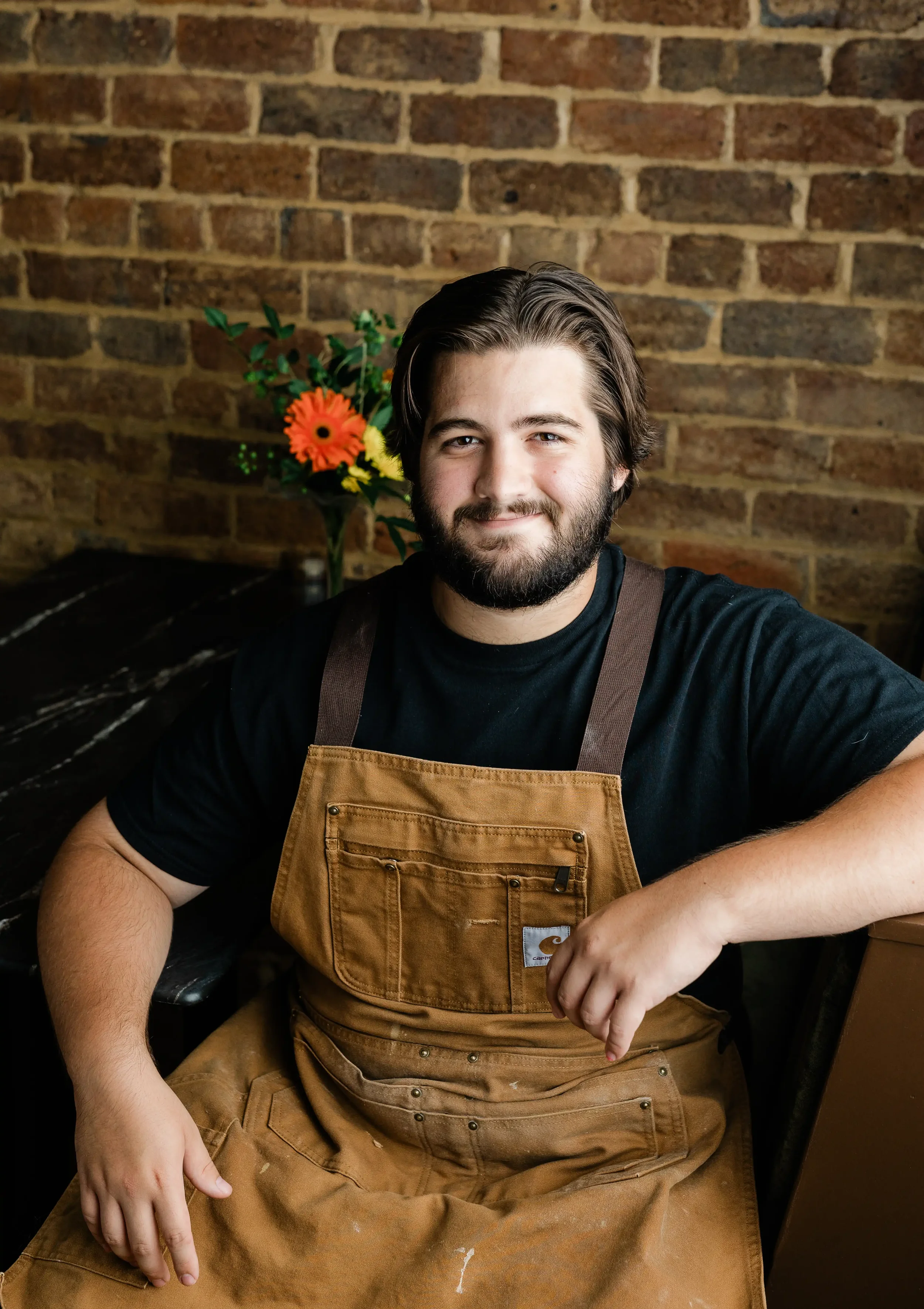 A man with a beard and dark hair, wearing a black shirt and a brown apron, takes a selfie in front of a brick wall. A bouquet of orange, yellow, and green flowers is in the background.