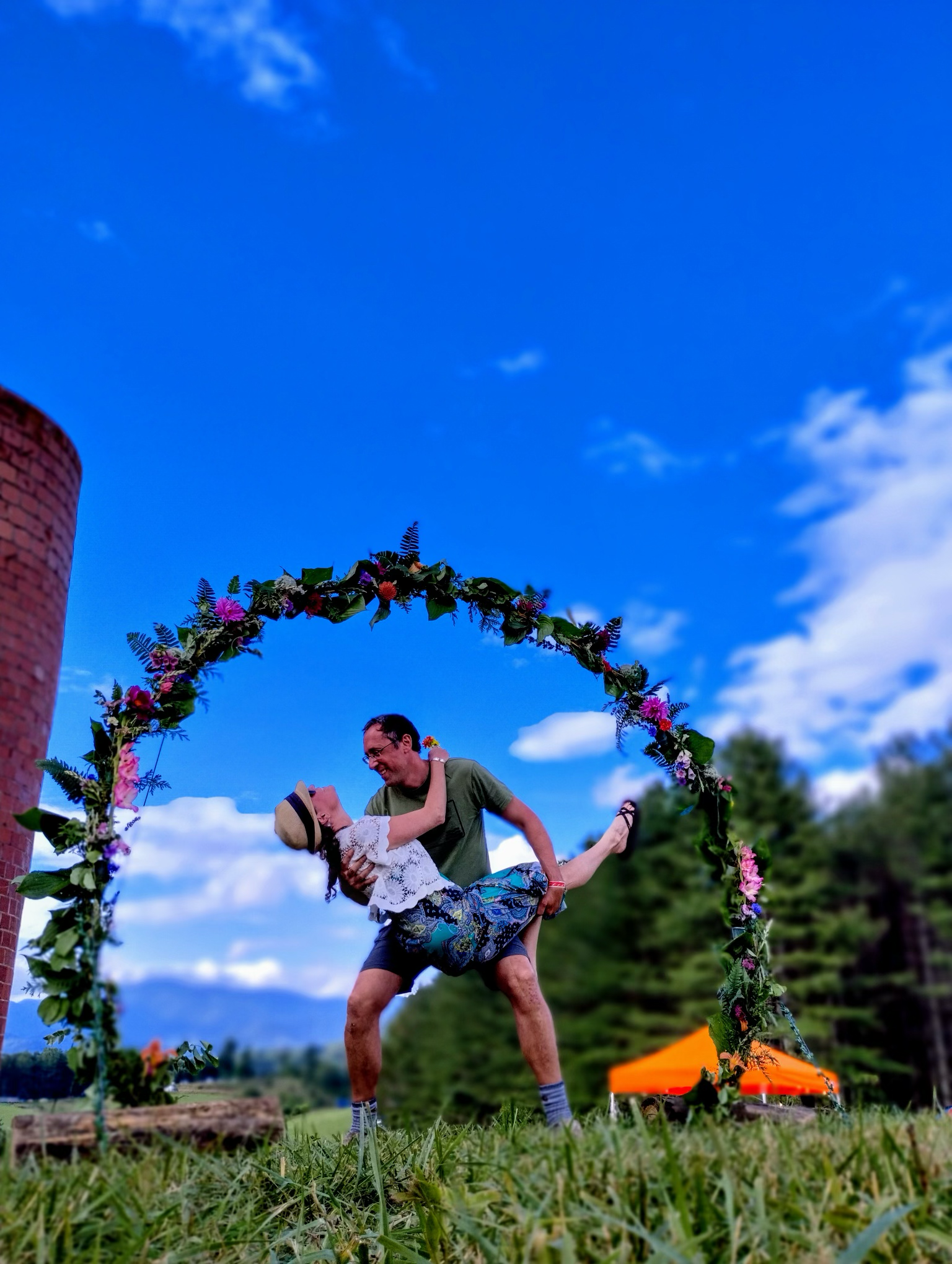 A man lifting a woman during a dance at an outdoor event, with a decorated floral arch and a partly cloudy sky in the background.