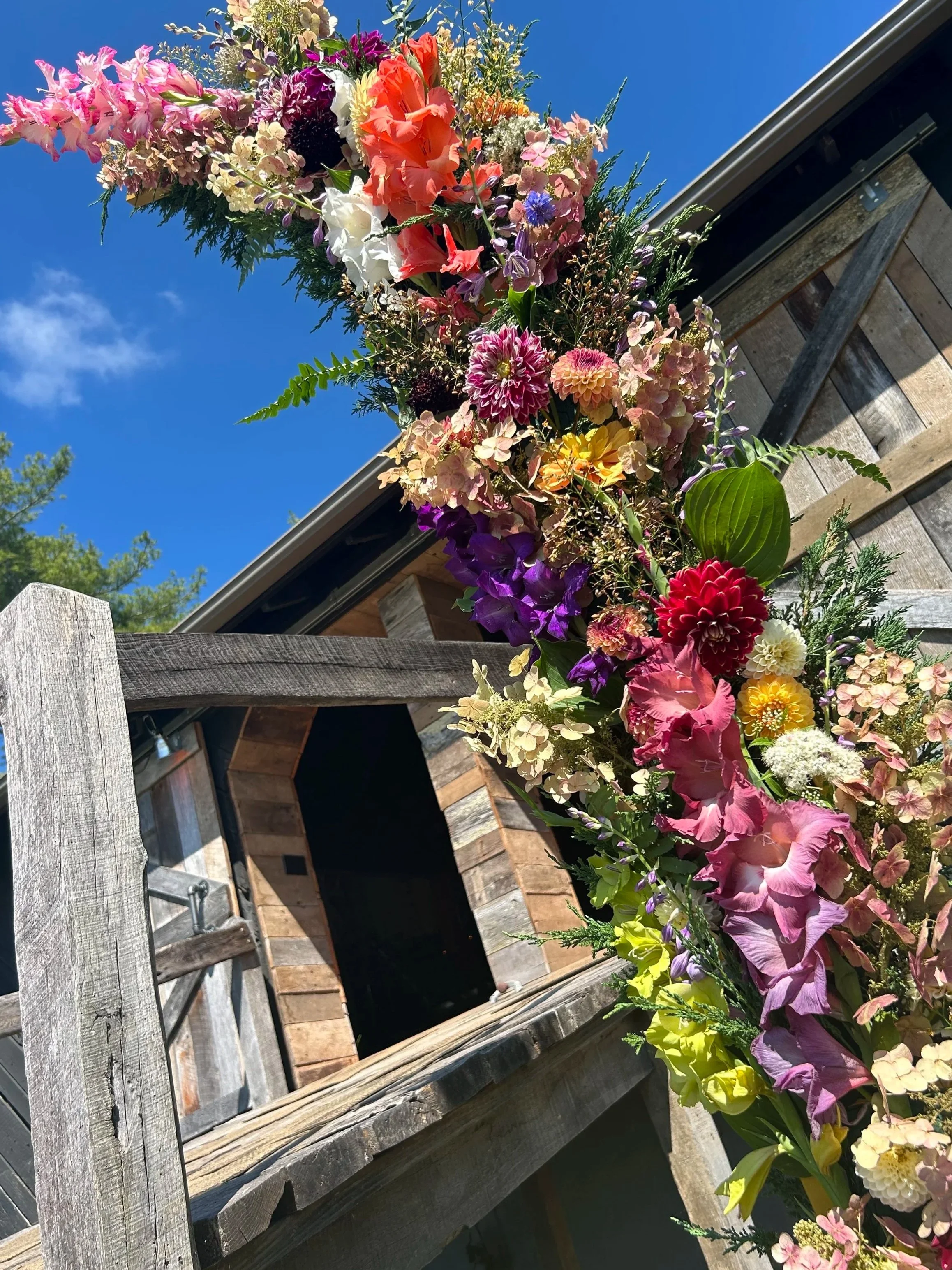 A colorful flower garland hanging on a rustic wooden fence against a bright blue sky.