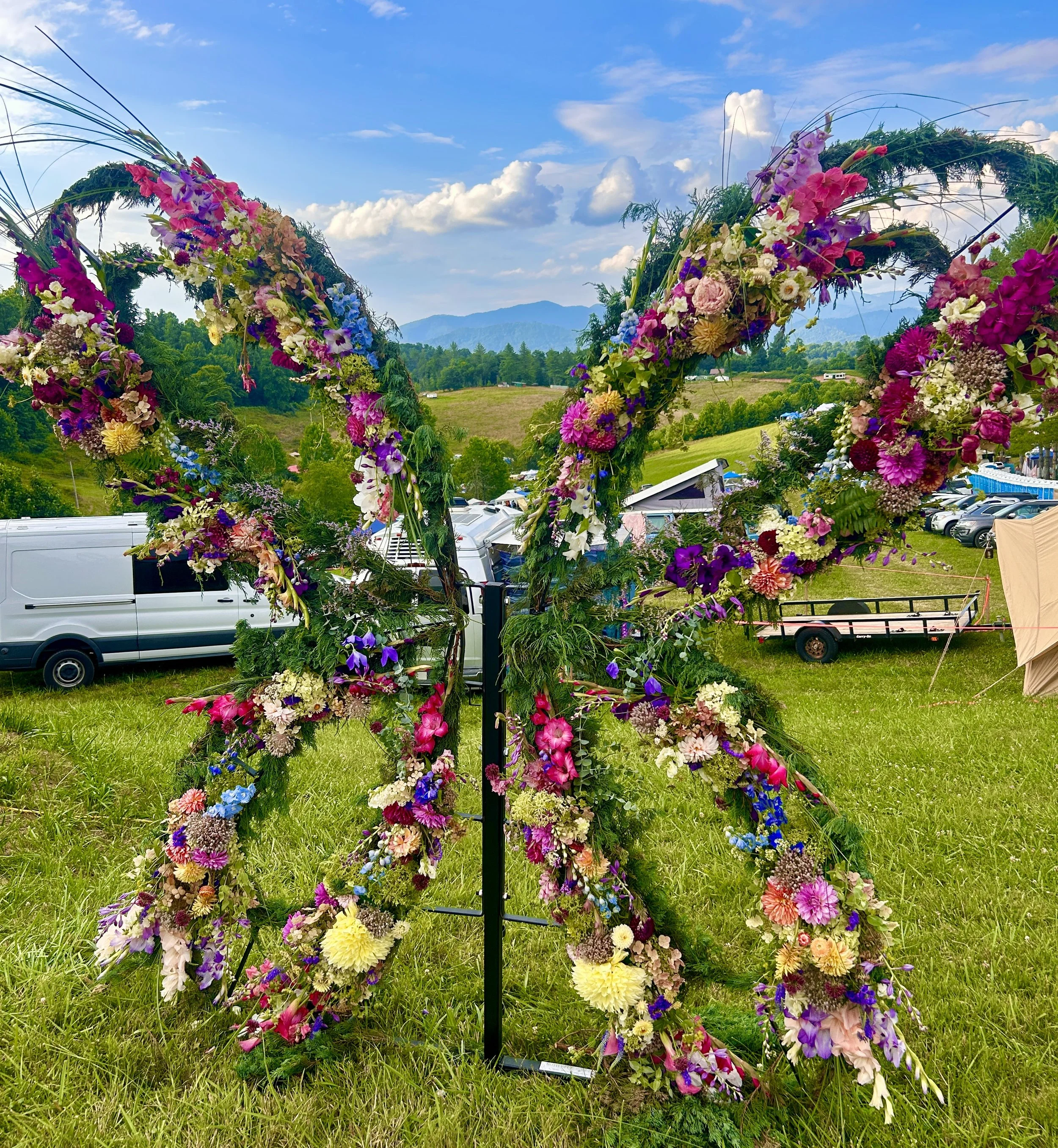 Colorful floral arrangement arch with various flowers in pink, purple, yellow, and white on a green grassy field, with a backdrop of trees, mountains, and a partly cloudy sky.