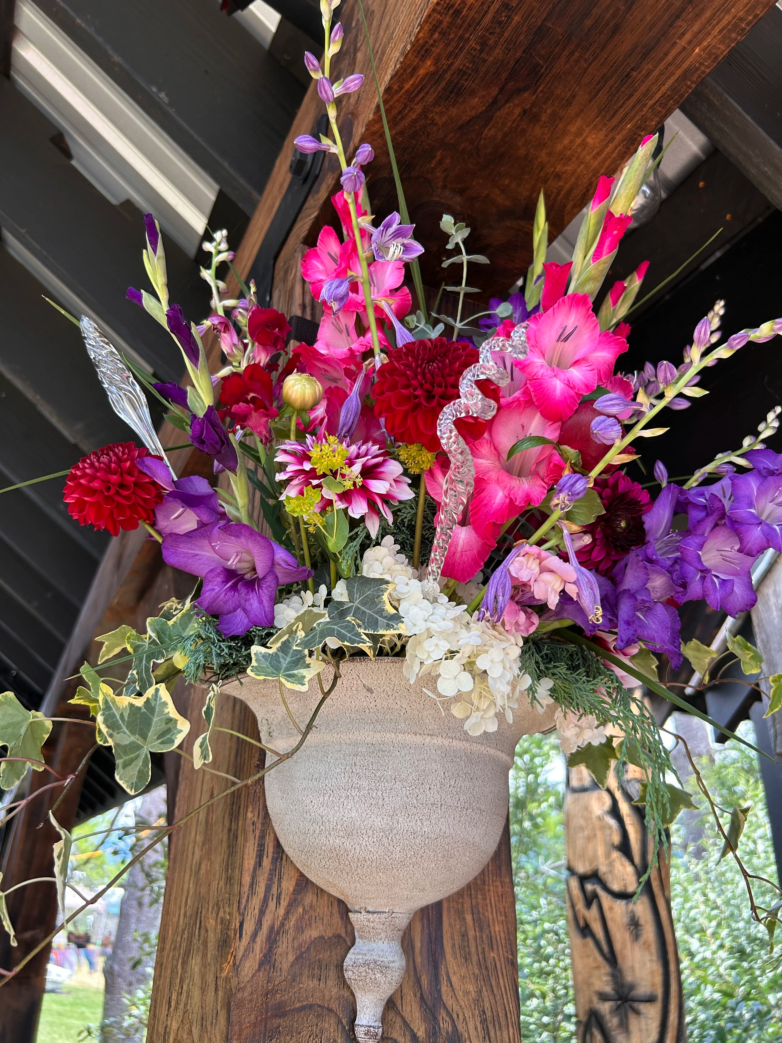 A vase filled with colorful flowers such as pink, purple, and red blooms, including gladiolus, dahlias, and other flowers, with green foliage and decorative elements, placed on a rustic wooden table.