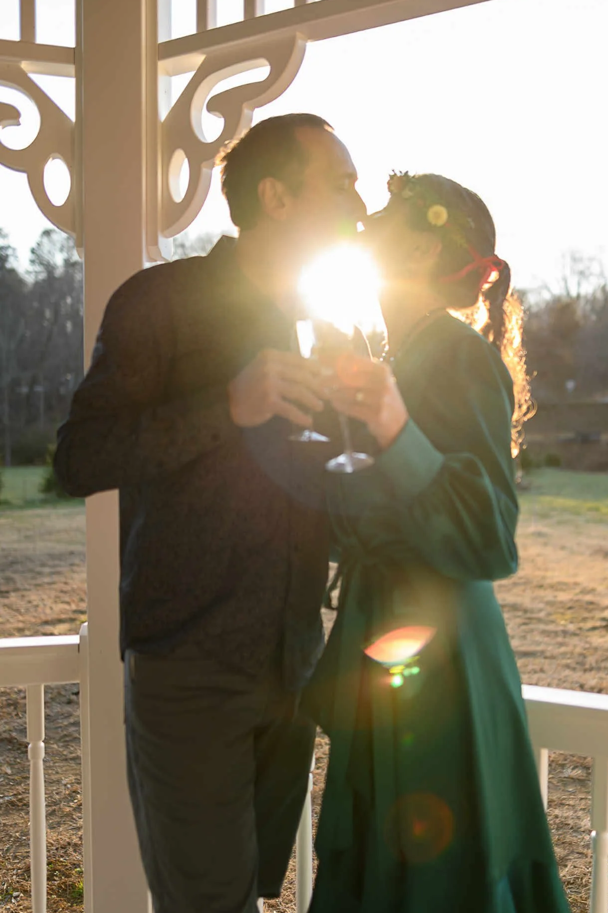 A man and woman sharing a kiss while holding wine glasses, backlit by sunlight, standing on a porch with decorative white railing.