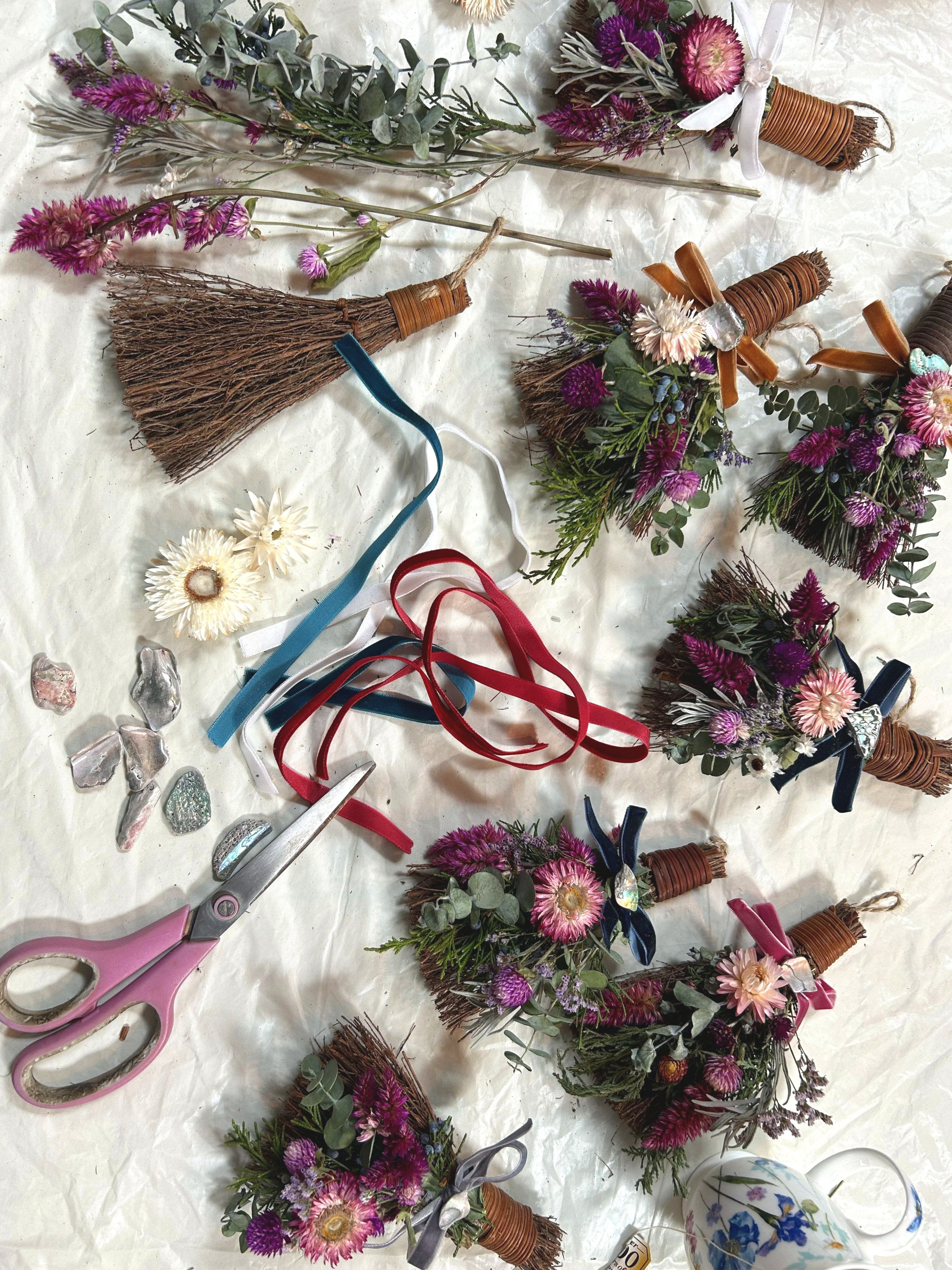 Various dried flower bouquets, some with ribbons tied around their stems, along with a pair of pink scissors, a ceramic teapot with blue floral patterns, and scattered small decorative stones on a white surface.