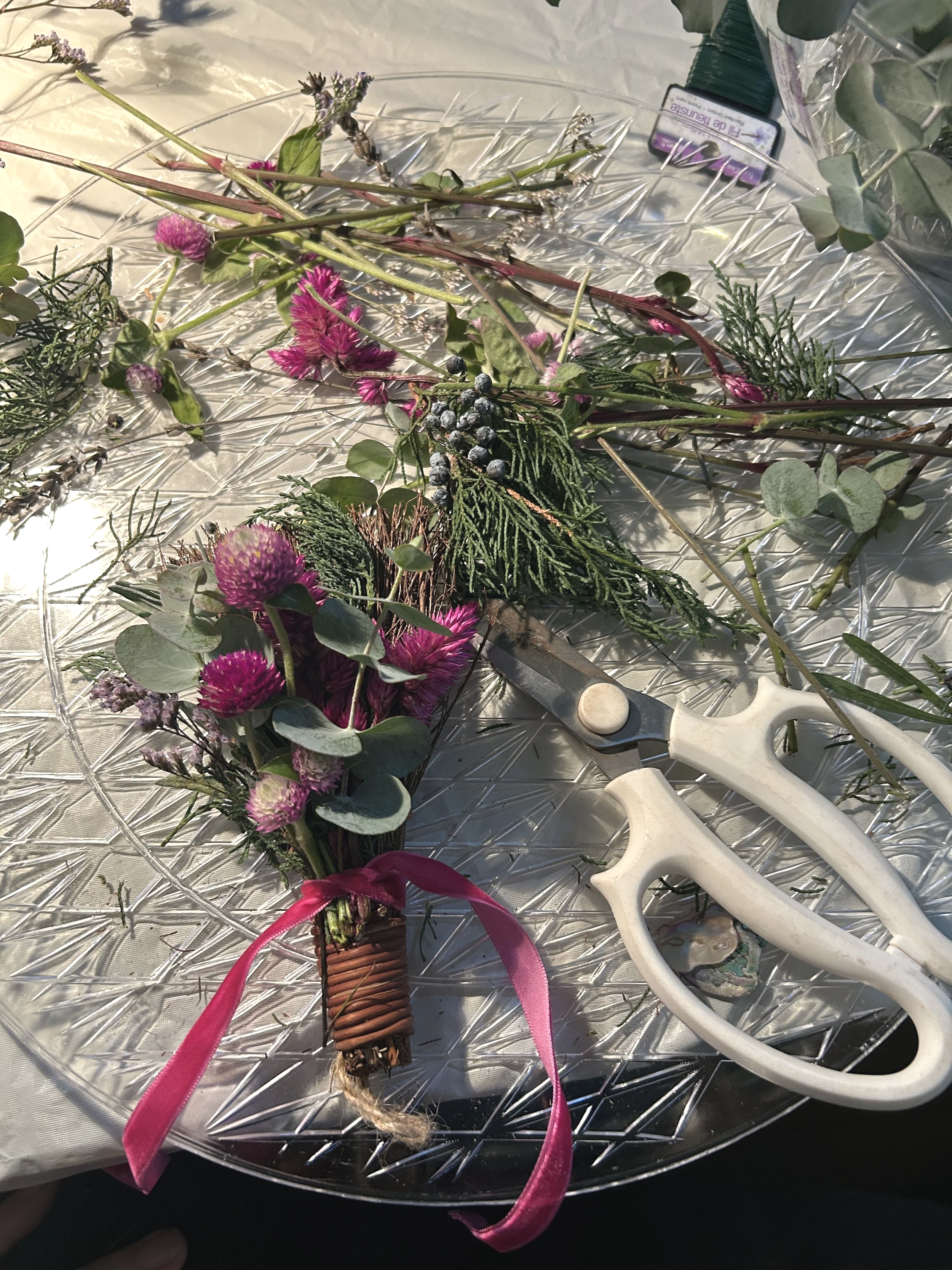 A bouquet of wildflowers with pink, purple, and green foliage wrapped with a pink ribbon on a glass tray with a candle snuffer and a pair of scissors nearby.