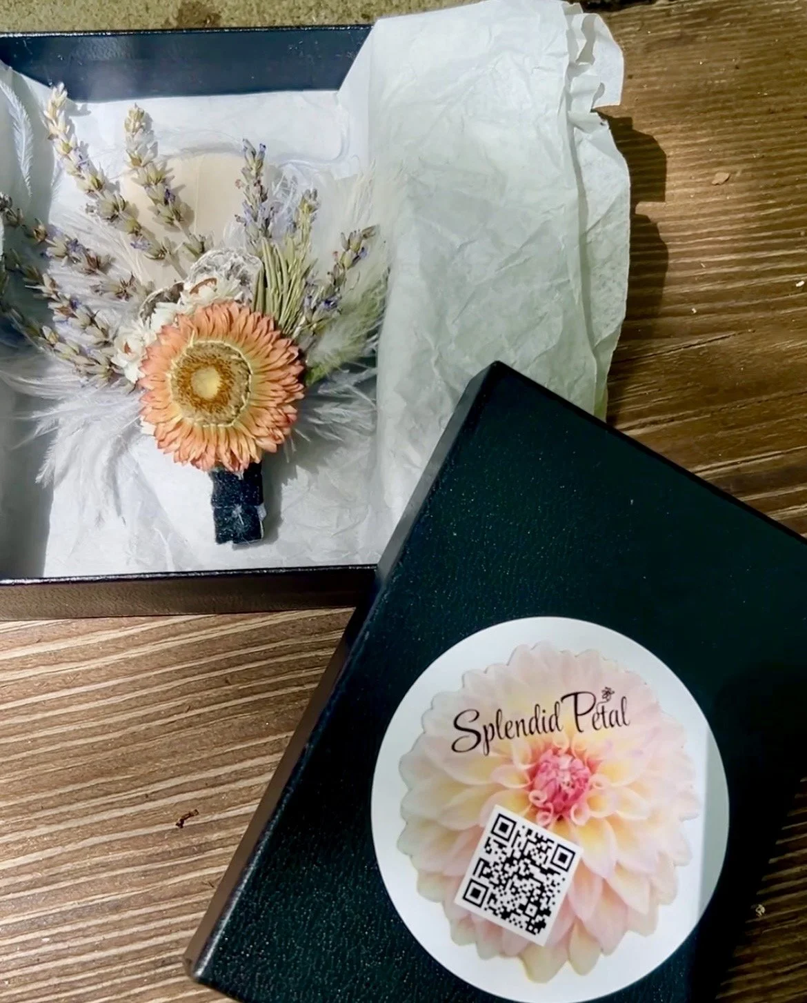 A bouquet of flowers with a large peach-colored Gerbera daisy, lavender sprigs, and white accents, placed inside a black gift box with tissue paper, and a black box with a 'Splendid Petal' sticker and QR code on a wooden surface.