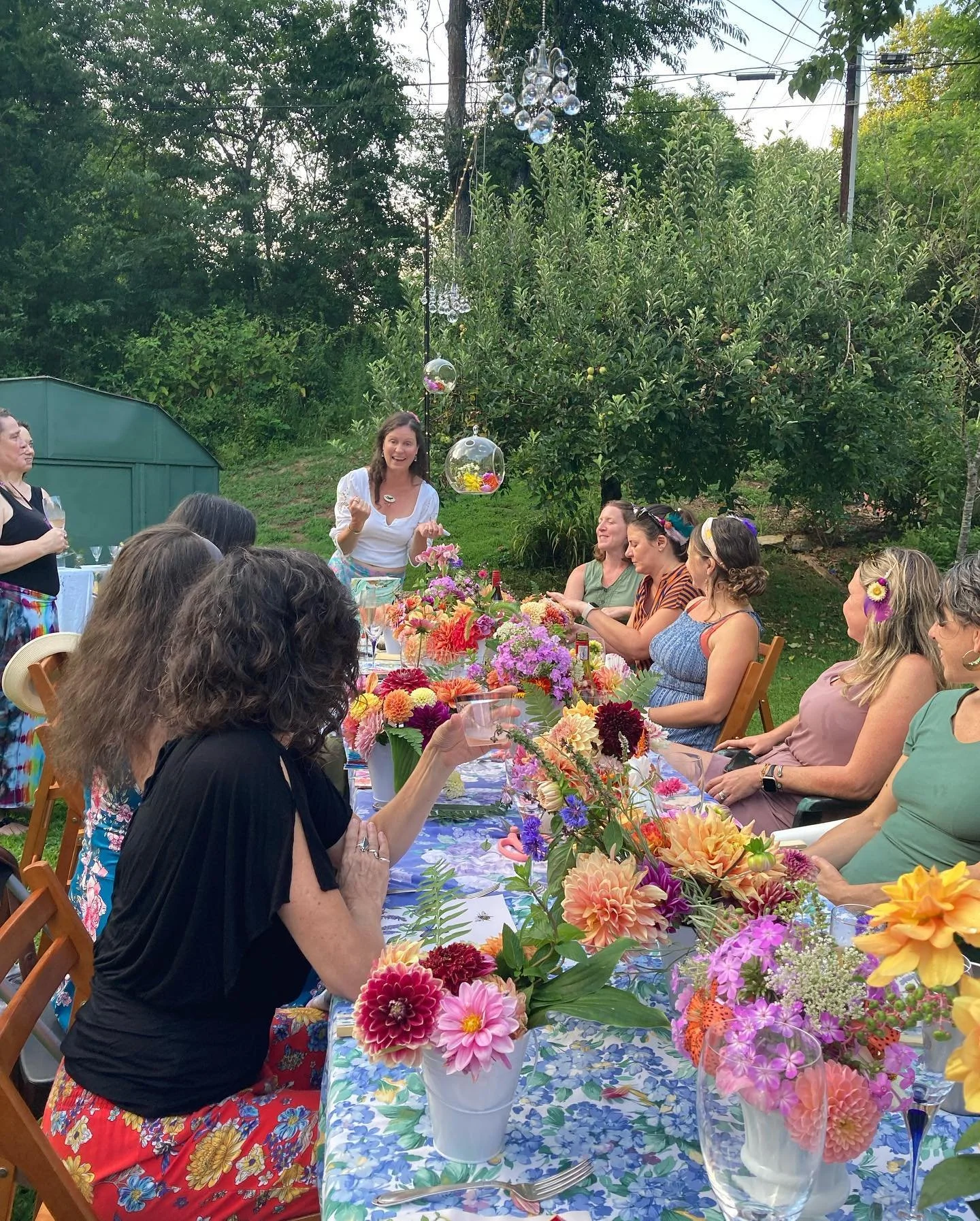 A group of women seated around a long outdoor table decorated with colorful flowers. One woman is standing and speaking to the group, with glass ornaments hanging above her. The setting appears to be a garden or backyard with trees and greenery in th