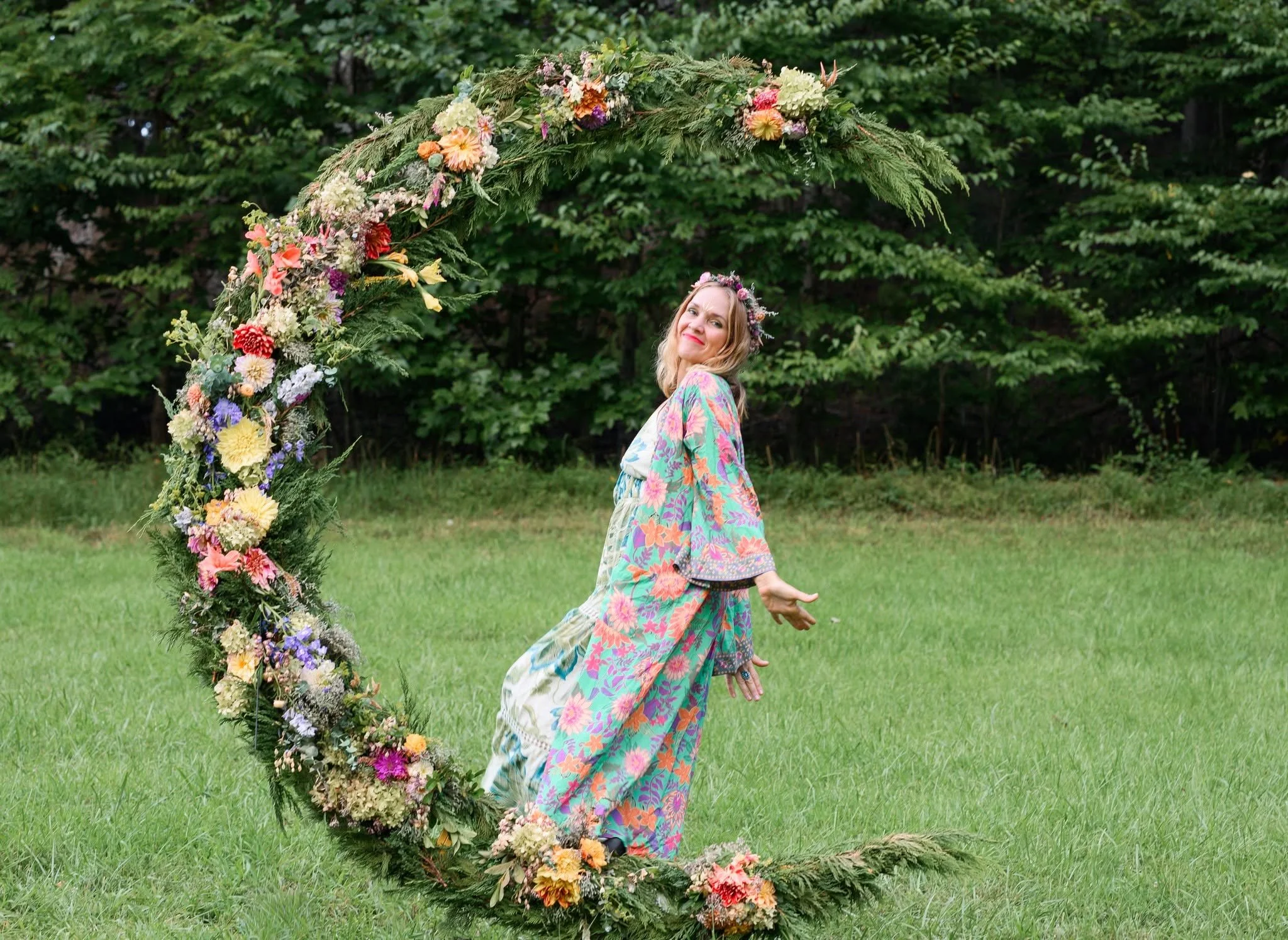 A woman with blonde hair and a floral crown, dressed in a colorful floral-patterned dress, standing behind a large, circular floral arch outdoors, with green trees and grass in the background.
