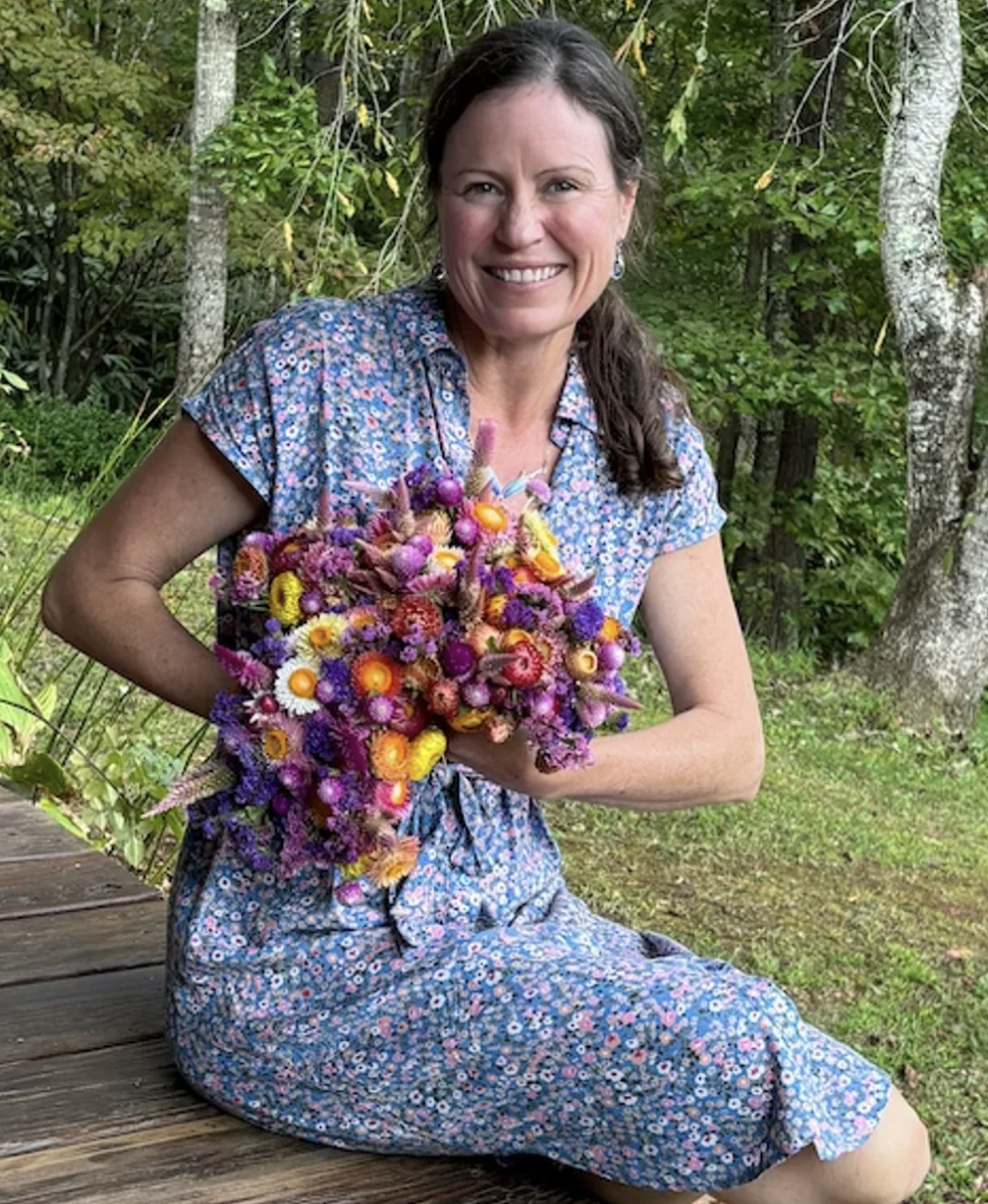 A woman with long dark hair sitting on a wooden surface outdoors, holding a large, colorful bouquet of flowers, surrounded by green trees and foliage.