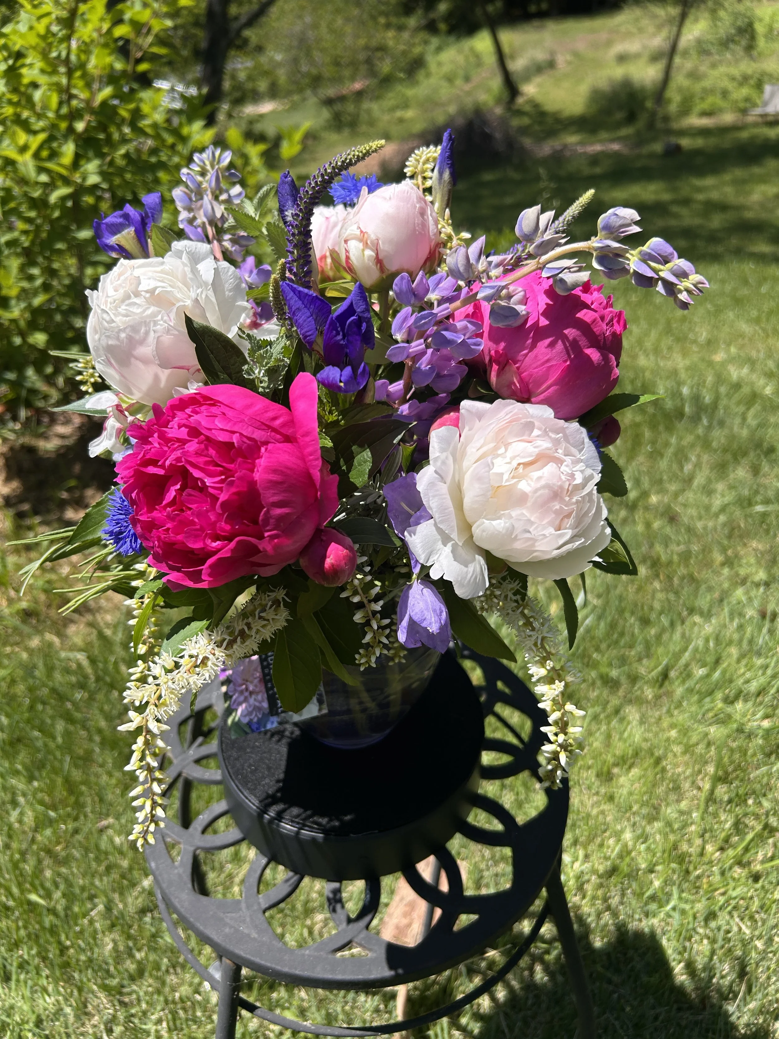 A vibrant bouquet of pink, white, purple, and blue flowers placed on a black metal outdoor table with a grassy yard and trees in the background.