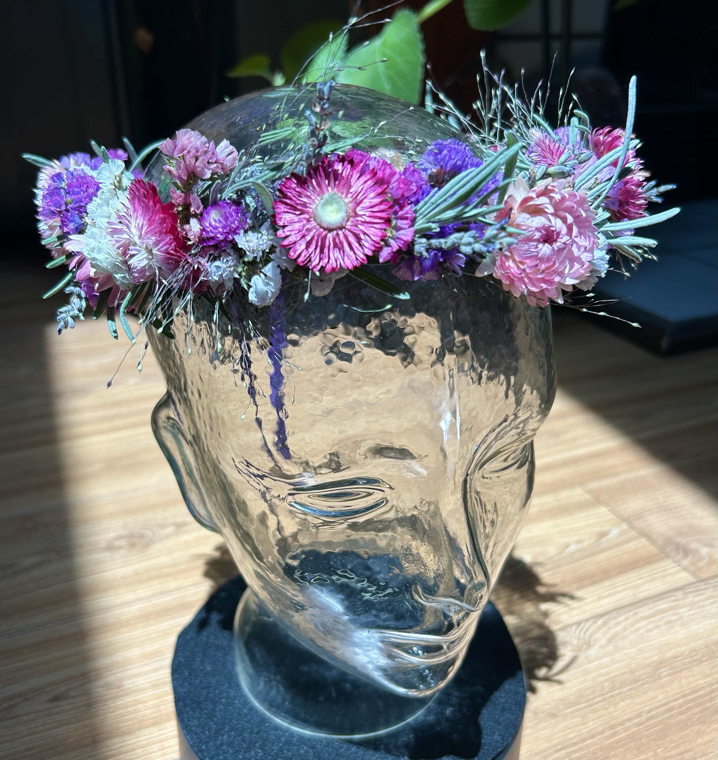 A clear glass vase with a floral arrangement of pink, purple, and white flowers, and greenery, sitting on a wooden surface, with a black stand or base visible underneath.