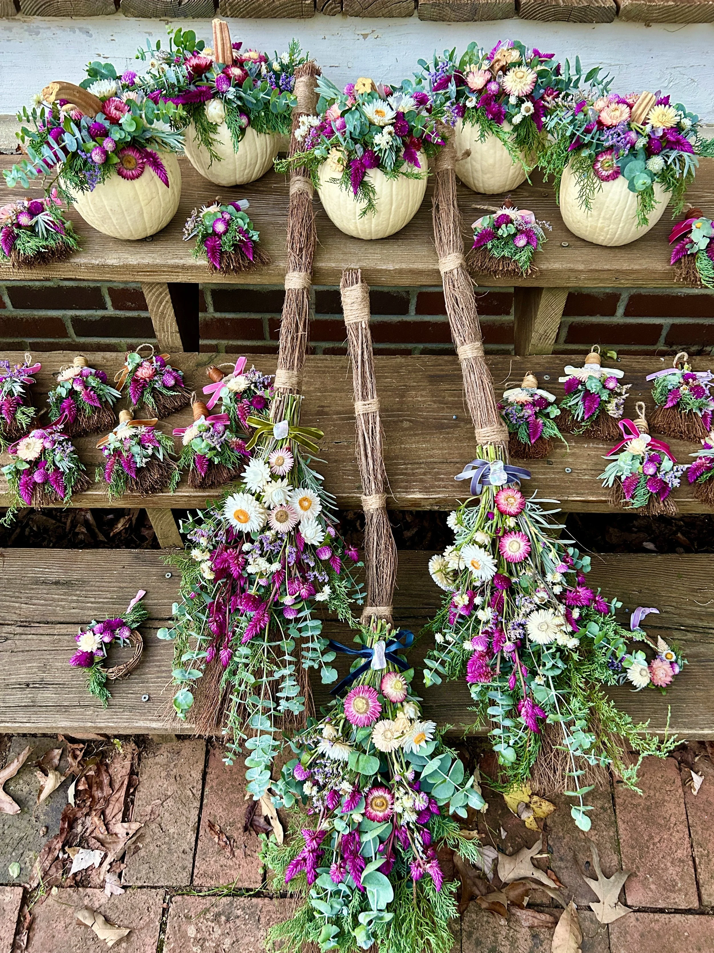 Colorful floral arrangements in pumpkin-shaped containers on a wooden bench, with some floral bouquets and greenery on the ground, against a brick and wood background.