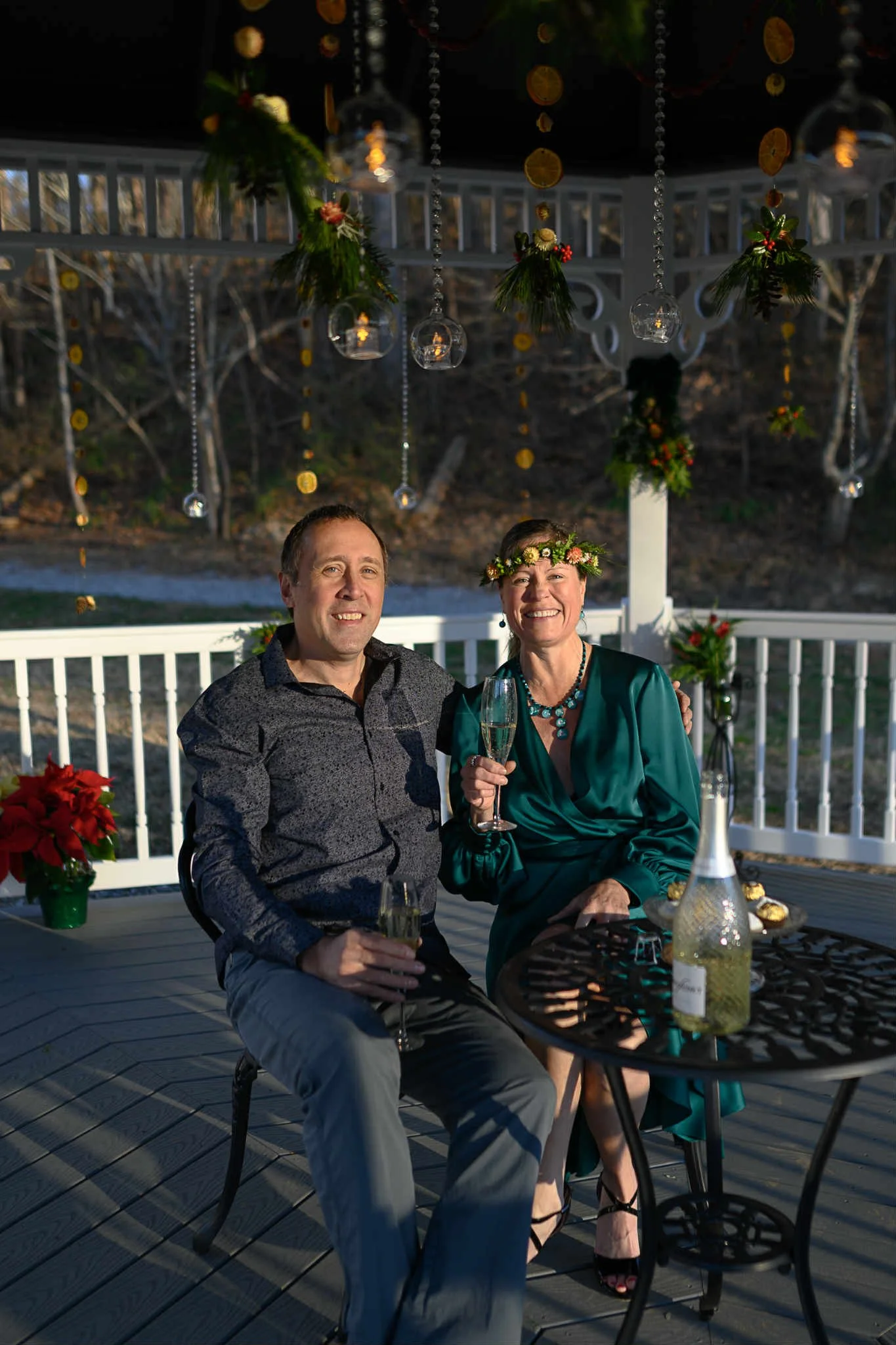 A man and woman celebrating at an outdoor party, dressed festively with holiday accessories, sitting at a table with drinks, surrounded by Christmas decorations and lights.