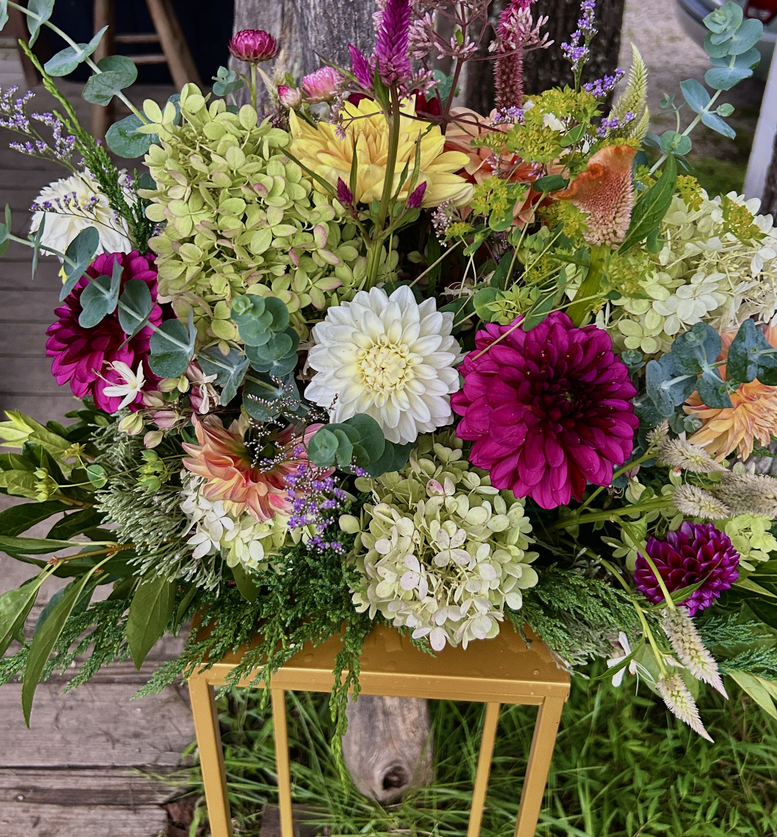 A vibrant bouquet of various flowers, including dahlias, hydrangeas, and other colorful blossoms, displayed on a small yellow table outdoors on a wooden deck.