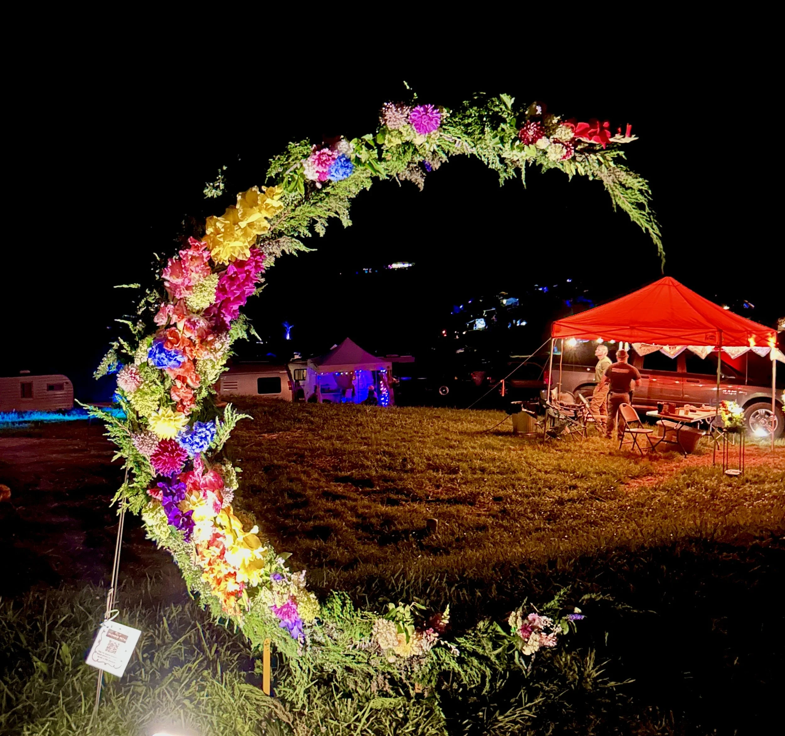 Nighttime outdoor scene with a colorful floral arch and people gathered under an orange canopy tent.