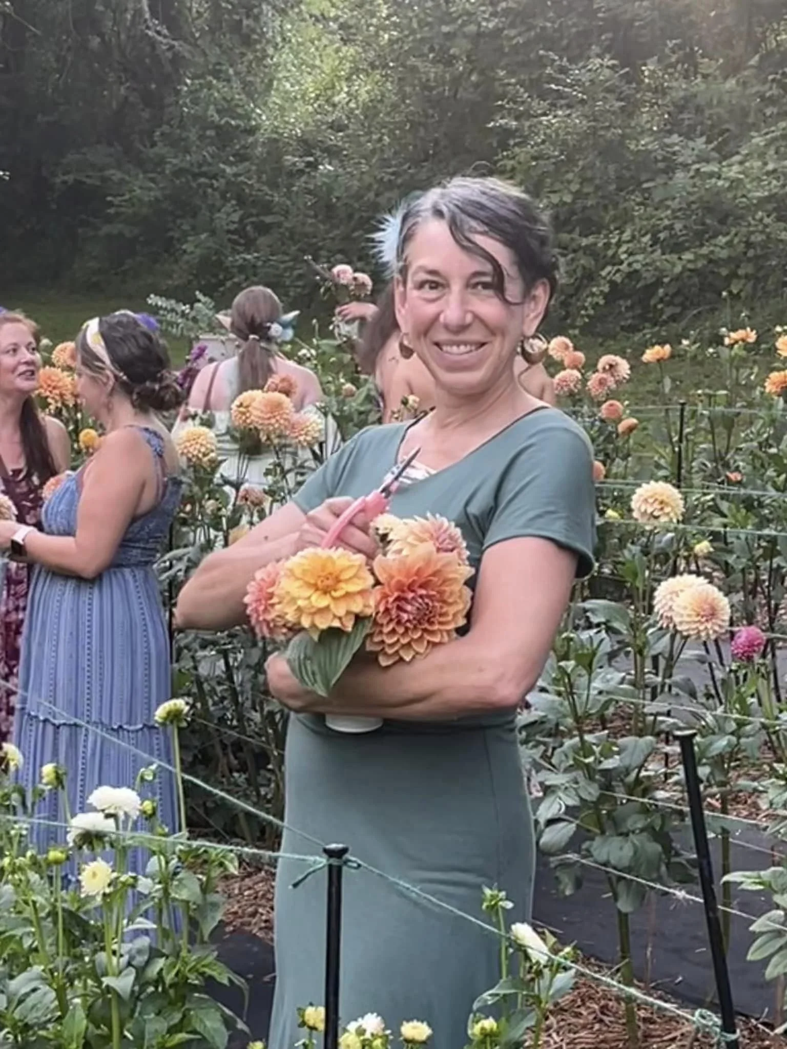 A woman with short gray hair and earrings standing in a garden with orange and yellow flowers, holding a bouquet of flowers and smiling at the camera.