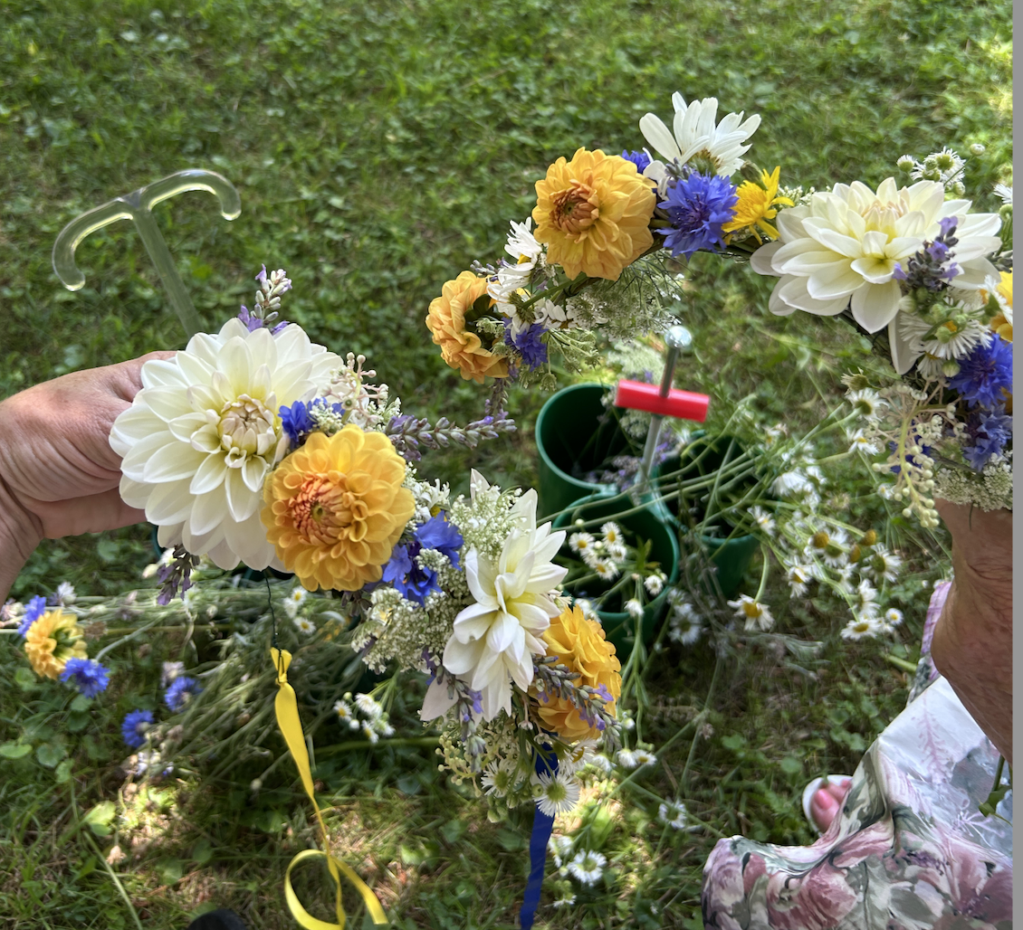 Person holding a flower crown made of various colorful flowers, including white dahlias, yellow marigolds, and blue cornflowers, with a garden background.