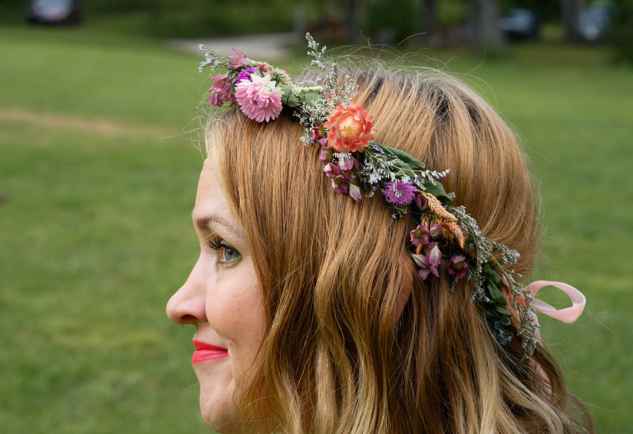 Side profile of a woman with red hair wearing a floral headband made of pink, orange, and purple flowers, outdoors with green grass and trees in the background.