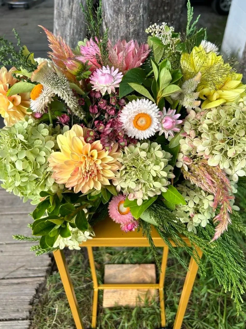 A colorful bouquet of mixed flowers including dahlias, daisies, hydrangeas, and greenery arranged in a yellow flower stand, placed on a wooden surface outdoors.